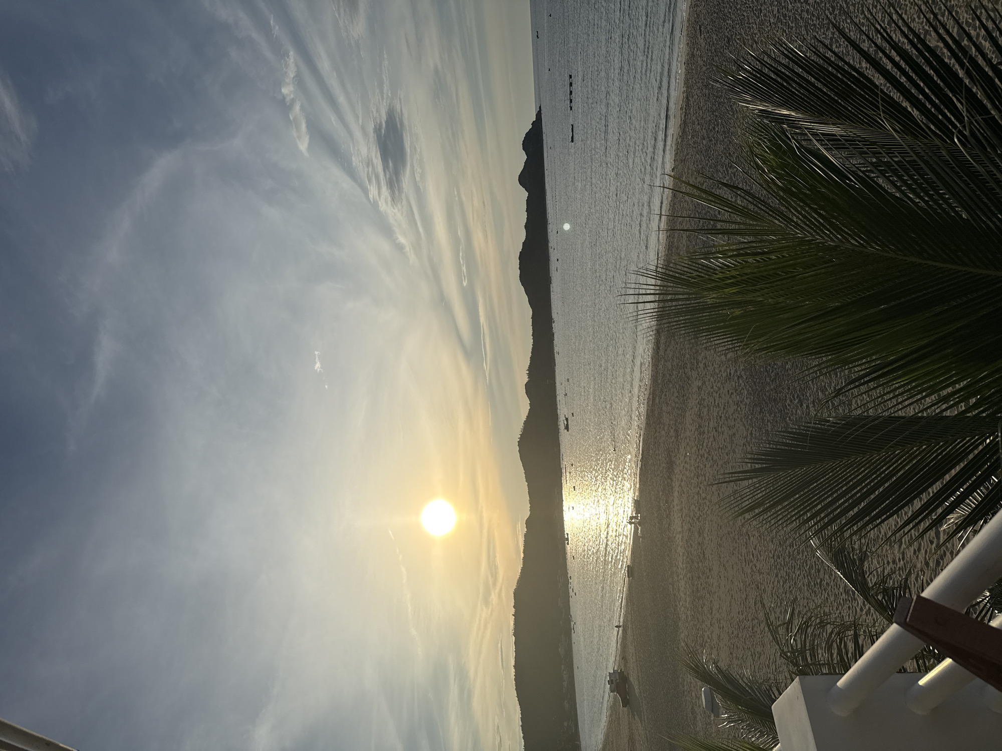 The image depicts a serene beach scene in Song Cau, Vietnam, during the late afternoon or early evening. The sun, a bright orb, is low on the horizon, casting a warm, golden glow across the sky and water. Wispy clouds streak across the blue sky, adding texture and depth.
In the foreground, lush green palm fronds are visible, framing the view of the beach and ocean. The sandy beach stretches along the coastline, appearing mostly empty except for a few scattered individuals and a parked vehicle on the left side. The ocean is calm, with gentle waves lapping the shore. Several small boats are visible scattered across the water, some anchored near the shore and others further out.
In the distance, rolling hills form a picturesque backdrop to the bay. The silhouette of these hills is softened by the descending sun. The overall atmosphere is peaceful and tranquil, evoking a sense of relaxation and natural beauty.