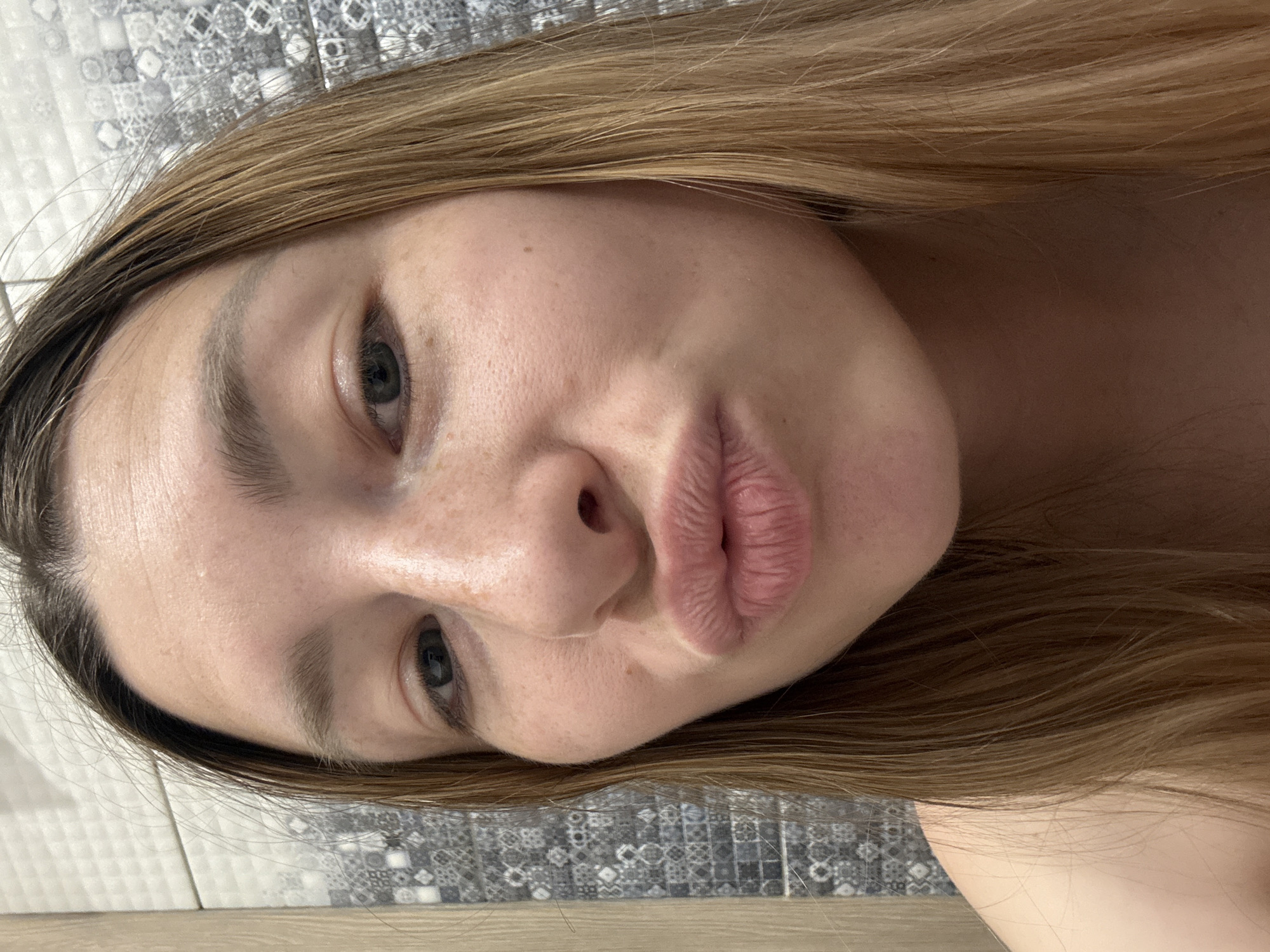 A close-up, angled portrait of a woman with light brown hair and fair skin. Her eyes are blue-gray. She has full lips and is making a slight pouting expression. Her skin appears clear with some freckles. The background consists of a tiled surface with a geometric pattern. The location is not discernible from the provided image.
