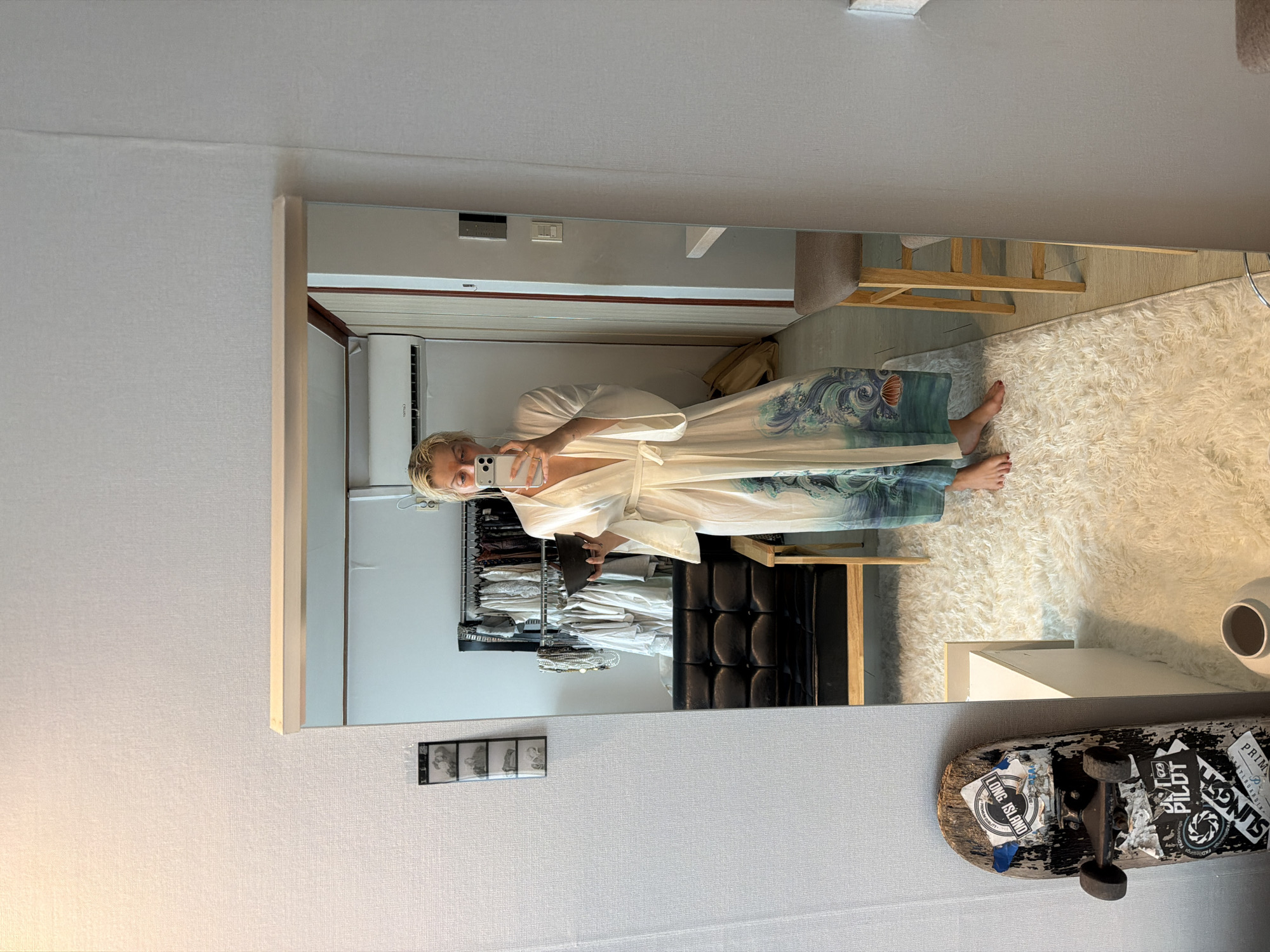 A Caucasian woman is photographed in a mirror selfie in what appears to be a modern apartment in Seoul, South Korea, as indicated by the presence of Korean power outlets. She is wearing a white robe with a blue wave design and holding a smartphone in her right hand to take the photo. Behind her is a walk-in closet with clothes on racks and a black leather chair. In the reflection, a beige chair, a white rug, and a white vase are visible. Below the mirror, there is a skateboard and a strip of what appears to be negatives or photographic film. The room has white walls and the lighting seems bright.