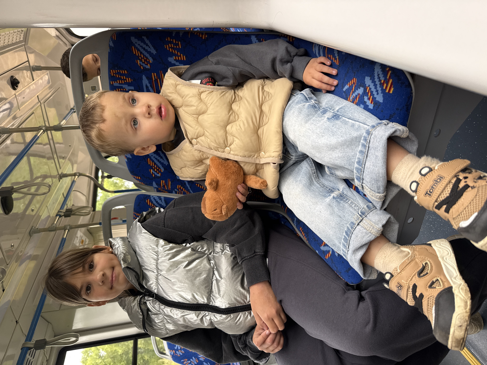Two children are seated on a bus. On the left, a girl with brown hair, wearing a silver puffer vest over a dark sweatshirt and dark pants, looks at the camera. Next to her, on the right, a young boy with blonde hair and blue eyes, wearing a light tan puffer vest over a dark sweatshirt and light blue jeans, holds a small, light brown stuffed animal. The boy is looking away from the camera. The bus seats are blue with a red and yellow pattern. A third child, partially visible, is seated behind them. The bus appears to be in Rusanovo, Russia.