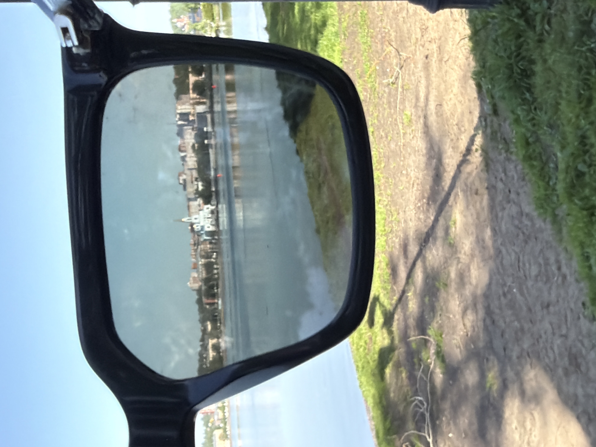 The image depicts a pair of sunglasses held up, with the reflection in the lenses showing a cityscape across a body of water. The cityscape appears to be Votkinsk, Russia, judging by the visible architecture resembling the Cathedral of the Annunciation and other buildings along the waterfront. Outside the frame of the sunglasses, the immediate environment shows a dirt path with grass on either side, suggesting a park or natural area. The scene is captured during daylight hours under a clear sky. The positioning of the sunglasses allows the viewer to see both the reflected landscape and the immediate surroundings.