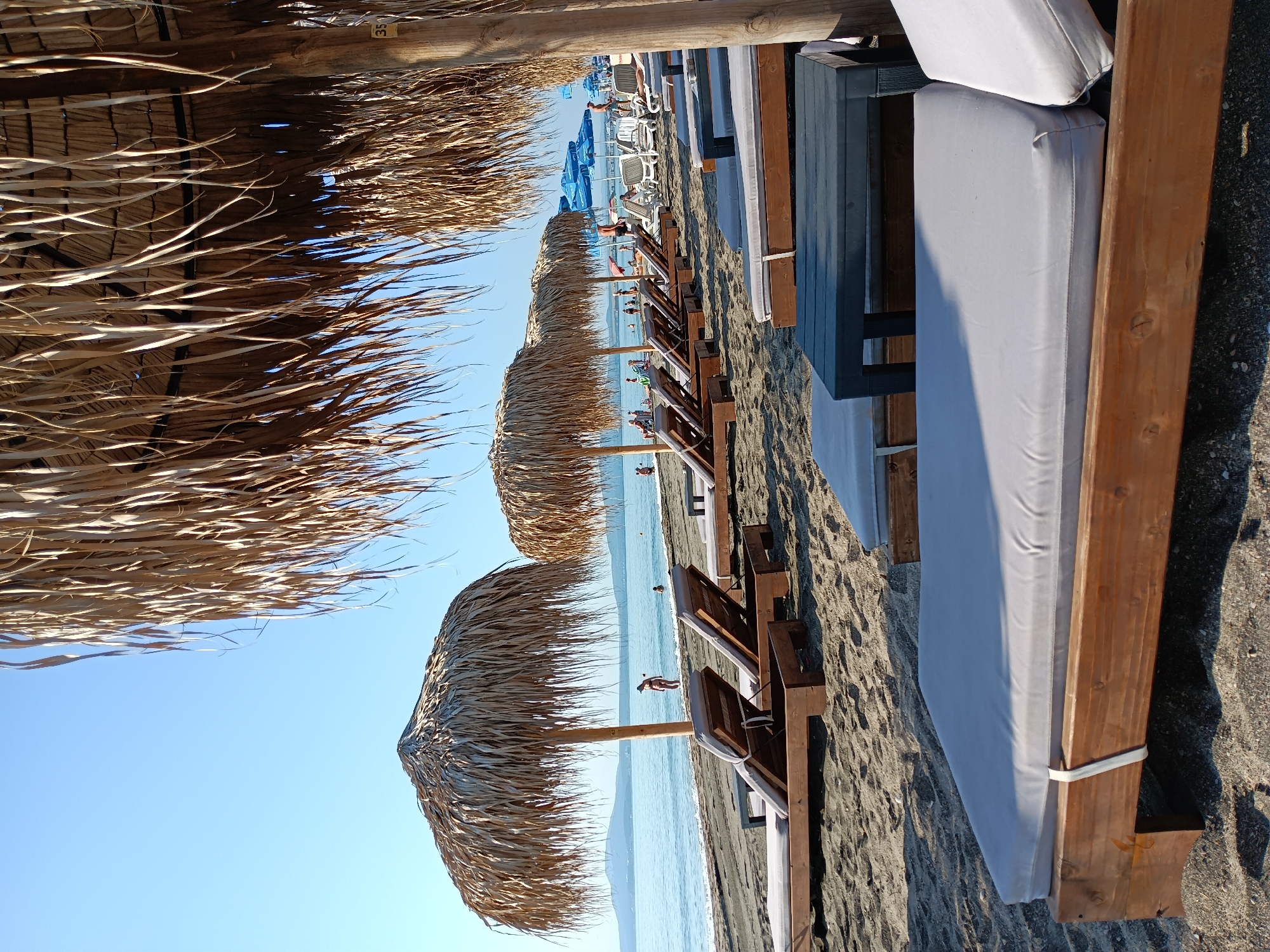 The image depicts a beach scene with rows of empty lounge chairs and thatched umbrellas under a clear blue sky. The beach is composed of dark sand and pebbles, with the sea visible in the background. There are a few people in the shallow water and scattered across the beach, some appearing to be sunbathing or walking. The lounge chairs are made of wood with light gray cushions. In the foreground, a dark rectangular table sits in front of a lounge chair. The overall impression is a tranquil and sunny day at the beach. This scene is likely from the beach in the city of Burgas, Bulgaria, which is known for its sandy shores and seaside resorts.