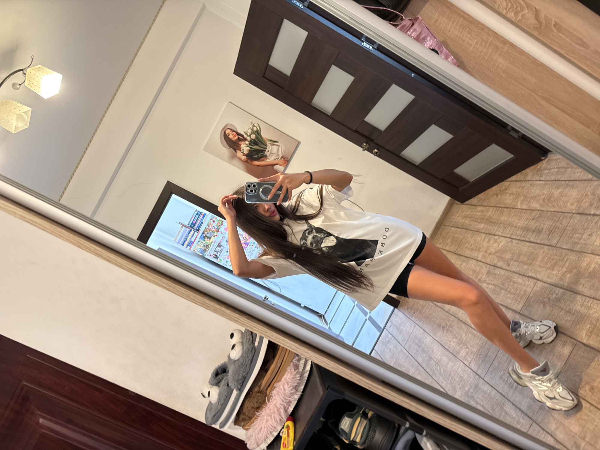 In a domestic indoor setting, a young woman with long dark hair is taking a mirror selfie. She is wearing a white oversized t-shirt with a black graphic of a dog, and black shorts. She is also wearing grey and white athletic shoes. The room has wood-paneled flooring and a dark brown door with frosted glass panels. A large mirror reflects the woman and part of the room. To the left, a shelf unit is visible with several pairs of slippers, including a pair of grey fluffy slippers with googly eyes, and a pink fuzzy slipper. A painting of a woman holding tulips hangs on the wall. The lighting appears to be from ceiling fixtures. The presence of indoor slippers and personal belongings suggests this is a residential interior, possibly a bedroom or hallway.