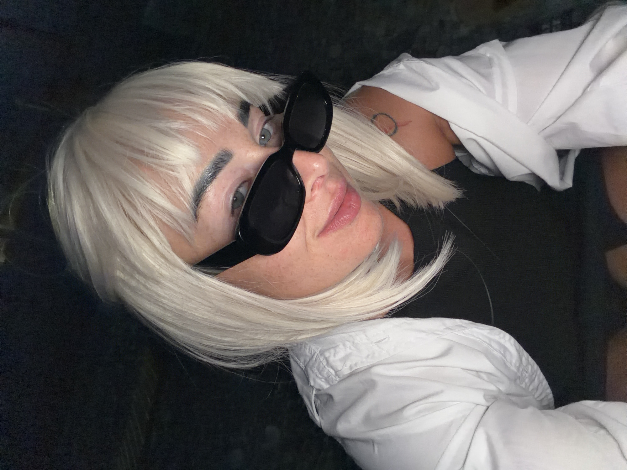 A woman with blonde hair, wearing black sunglasses and a white shirt, looks at the camera in an indoor setting. She has light skin, fair eyebrows, and light-colored eyes. A tattoo is visible on her left shoulder. The background is dark and out of focus. There is no indication of the city of Sochi, Russia, in the image.