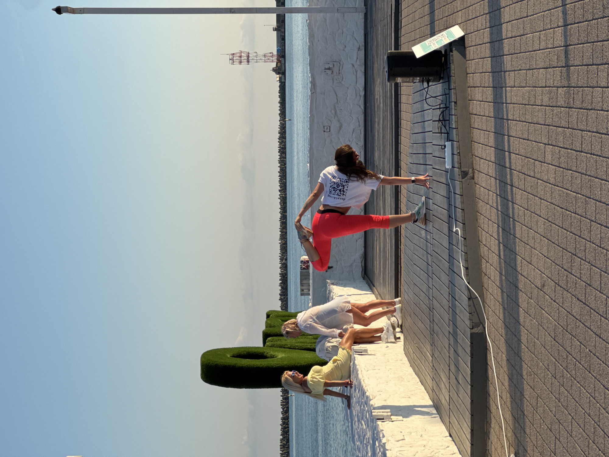 In Sochi, Russia, on a bright, clear day, three women are engaged in a yoga session outdoors. The scene is set on a paved walkway next to a body of water, likely the sea, with a breakwater visible in the distance. A large, green, sculpted bush resembling a number "0" is present on a white ledge.
One woman, dressed in bright red leggings and a white crop top, is in a dynamic split leap, showcasing her flexibility. She appears focused and graceful. Below her, two other women are in more grounded yoga poses. One wears a white dress and the other a yellow dress. They are positioned on the white ledge, seemingly participating in the same class or practice.
A notable detail is the text on the white crop top of the woman in mid-air. It features a QR code and the word "Инструктор" (Instructor) in Russian, indicating her role. A sign with a QR code is also visible on a pole near a speaker and electronic equipment, possibly for a class registration or information. The time of day appears to be late morning or early afternoon, judging by the sunlight casting shadows. The overall atmosphere suggests a healthy and active lifestyle being enjoyed by the participants in a scenic location.