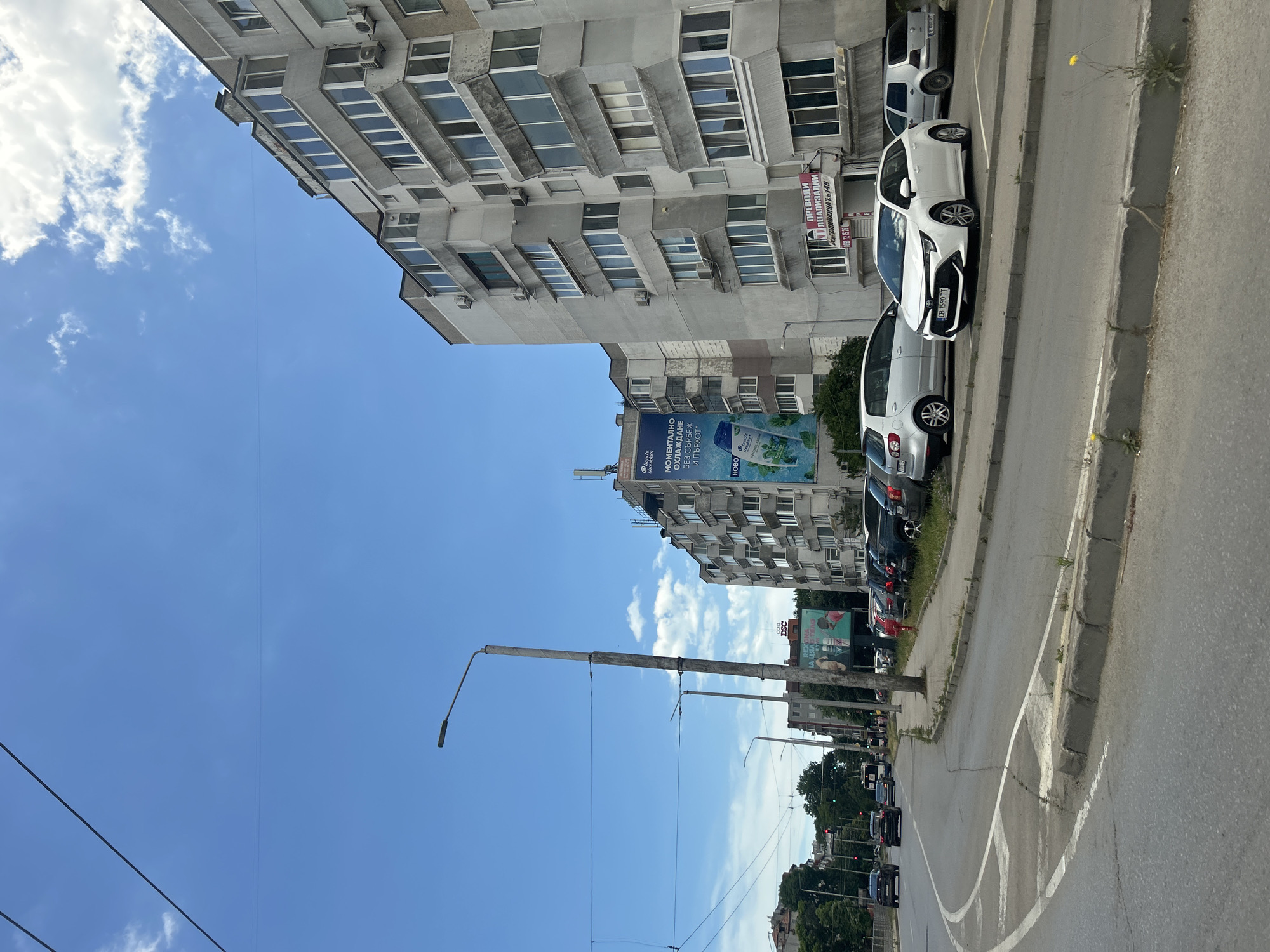 The image captures a daytime view of a street in Varna, Bulgaria, with a focus on a residential building and parked cars. The sky is predominantly blue with scattered white clouds. A tall street lamp stands prominently on the left side of the road, with power lines extending across the frame.
The residential building on the right is a multi-story structure, typical of Soviet-era architecture, with a light gray facade and numerous balconies. Some of the balconies appear to be enclosed with windows. Air conditioning units are visible on the exterior walls. There are commercial signs visible on the ground floor of the building, including one with red lettering above a glass door.
A row of parked cars lines the sidewalk in front of the building. Among them, a white sedan is parked in the foreground, its license plate clearly visible. Other vehicles, including a silver hatchback and a dark-colored SUV, are also parked.
The street in the foreground shows lane markings and a curb. The overall environment suggests an urban setting. No people are clearly visible in action, but a single figure in red is partially seen on the sidewalk further down the street.