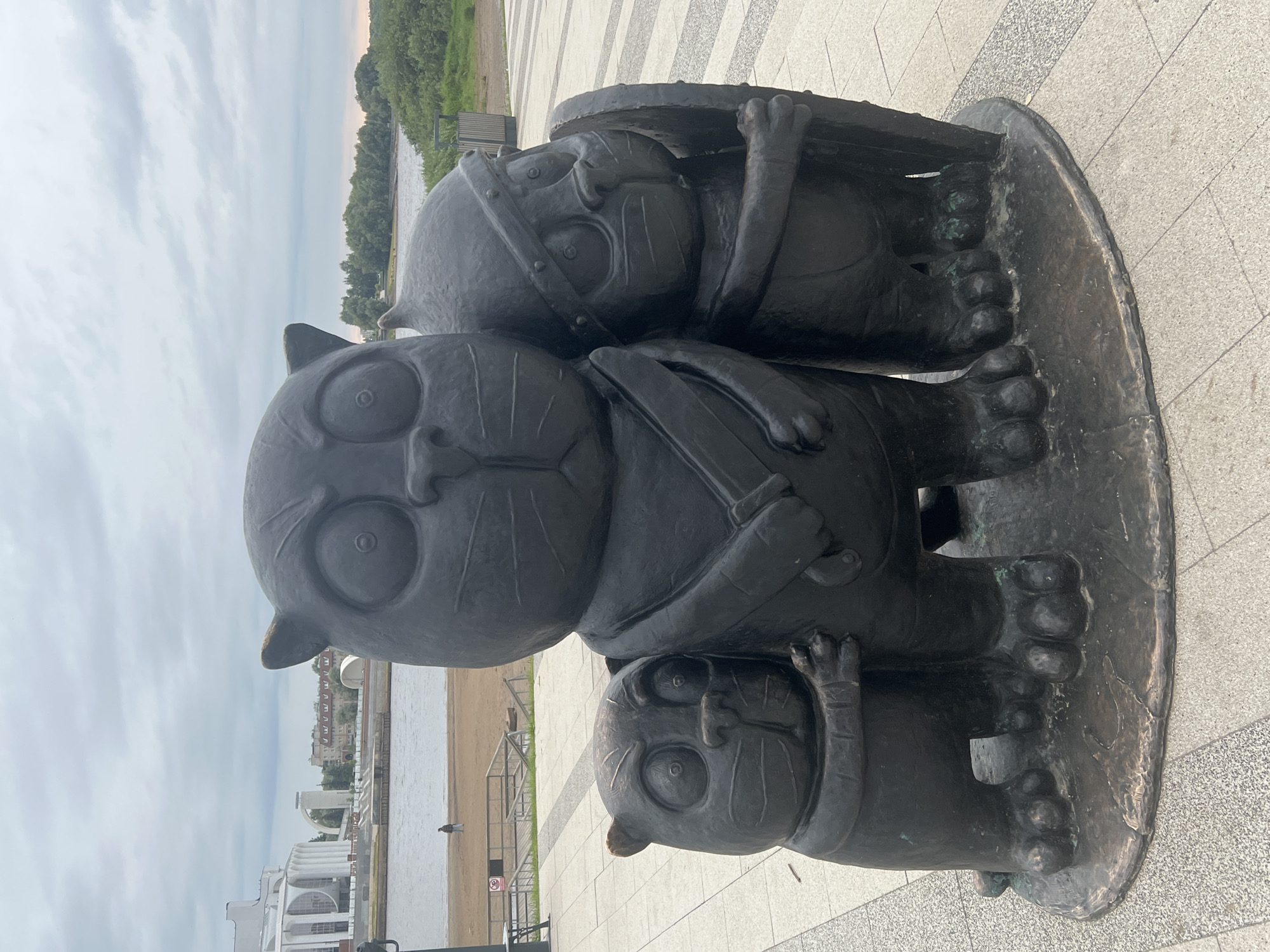 A bronze sculpture of three cartoonish cats stands on a paved promenade in Veliky Novgorod, Russia. The central cat is the largest, depicted with a stern expression, holding a sword across its chest. To its left is a smaller cat, embracing the central figure's leg, also with a wide-eyed, slightly apprehensive look. To the right of the central cat stands another cat, this one wearing a helmet and holding a shield, its gaze directed to the right. The sculpture is dark bronze and shows signs of age, with some greenish patina on the feet.
Behind the sculpture, a wide river flows, with a sandy embankment on the opposite side. A modern, white building with arched windows and a prominent dome-like structure is visible across the river, likely a cultural or public building. The sky is overcast, with soft grey clouds. The paved area in the foreground is laid with light-colored rectangular tiles. A single person is walking on the sandy embankment in the background, near a wooden barrier. The overall scene suggests an urban waterfront park or plaza.