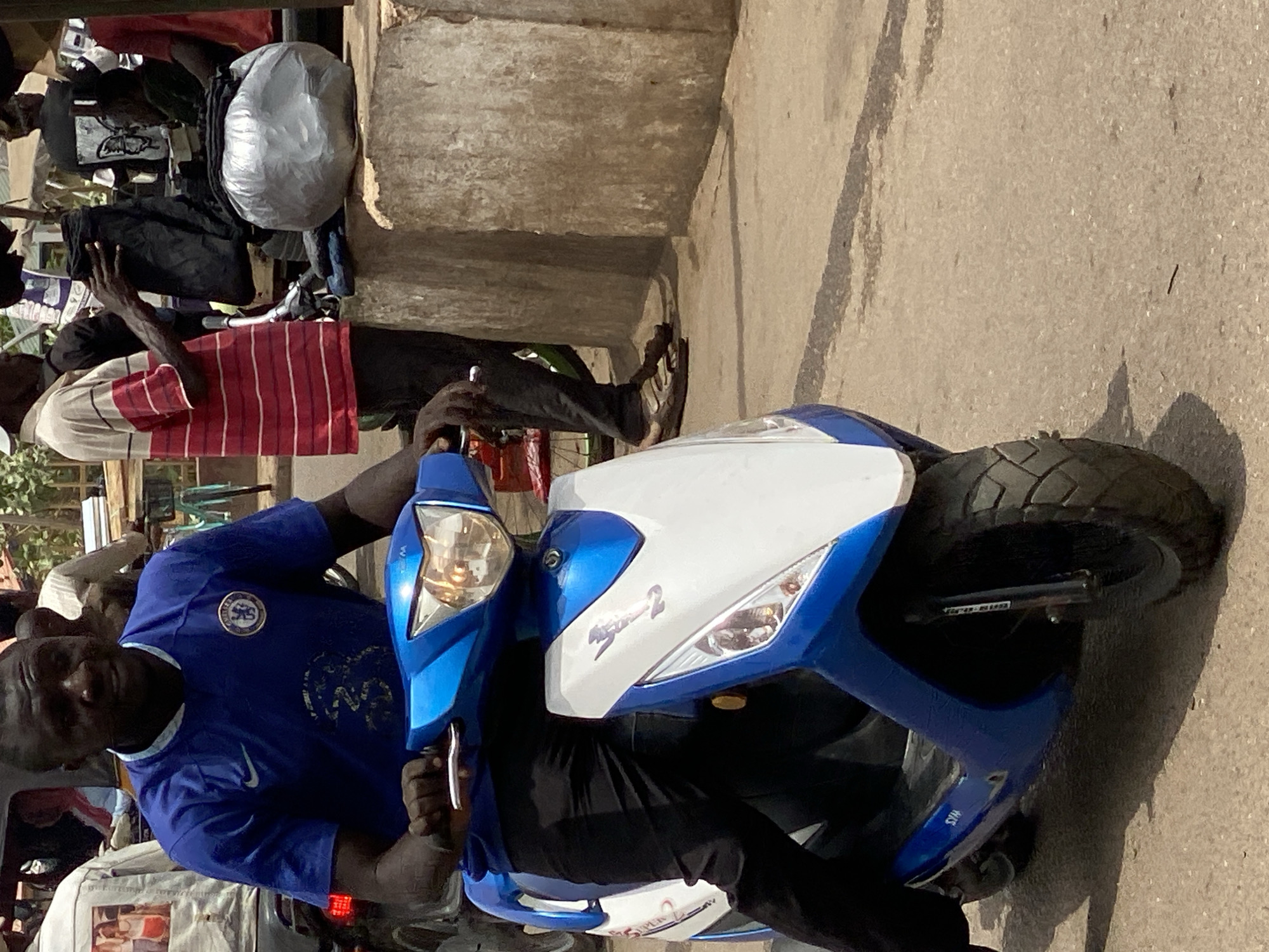 In Tudun Nupawa, Nigeria, a man wearing a blue Chelsea football club t-shirt sits on a blue and white scooter on a paved street. The man is looking at the camera. Several people stand behind the scooter, and objects like bags and clothes are visible. A concrete structure is to the side. The environment suggests a busy street scene.