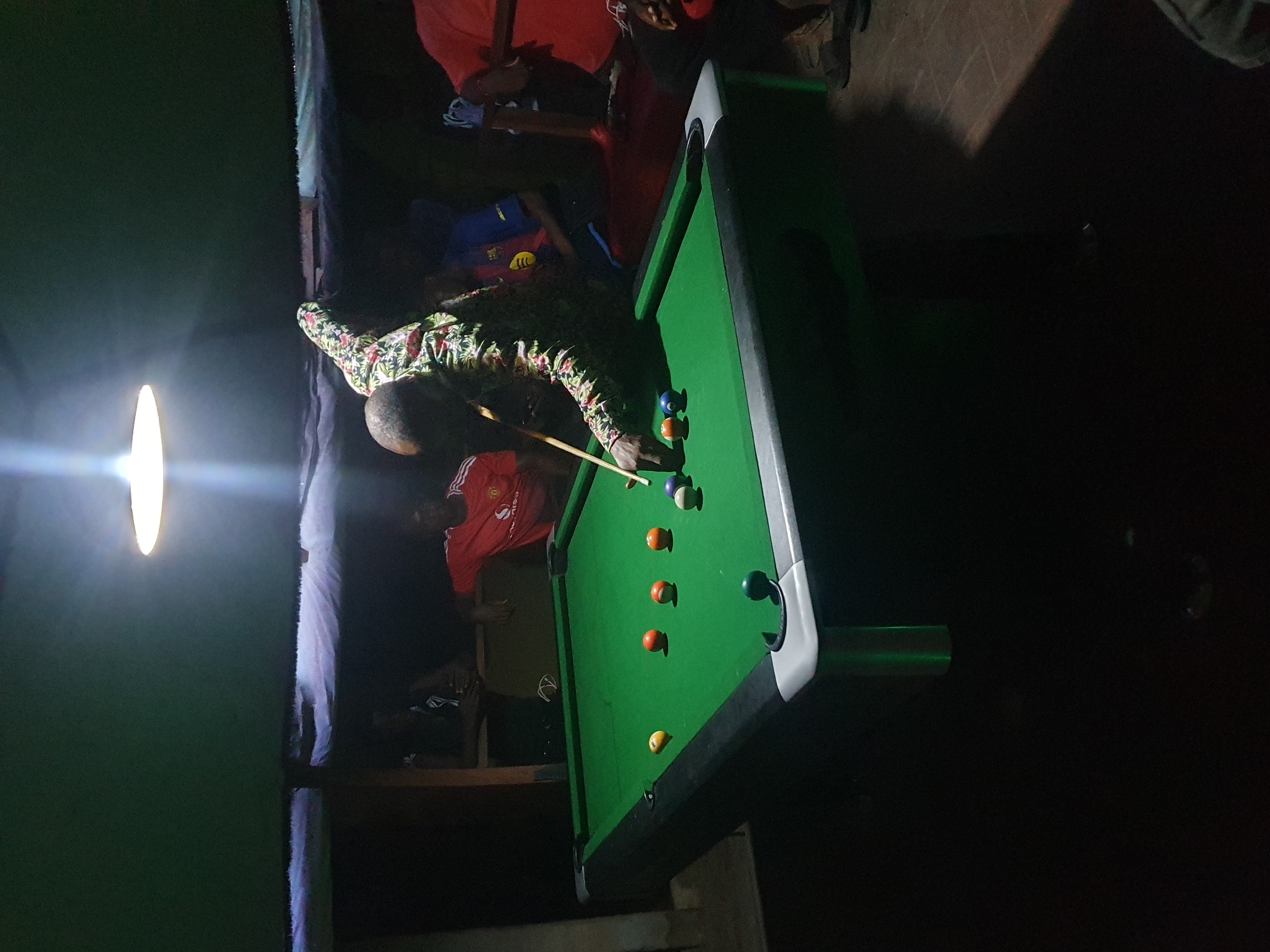 A brightly lit pool table dominates the scene in a darkened room in Lokoja, Nigeria. A man in a patterned shirt is positioned over the pool table, holding a cue stick, seemingly lining up a shot. Several pool balls are scattered on the green felt of the table. The room appears dimly lit except for a light source directly above the pool table. Other people are visible in the background, including one wearing a red Manchester United jersey. The environment seems to be a casual gaming or entertainment space.