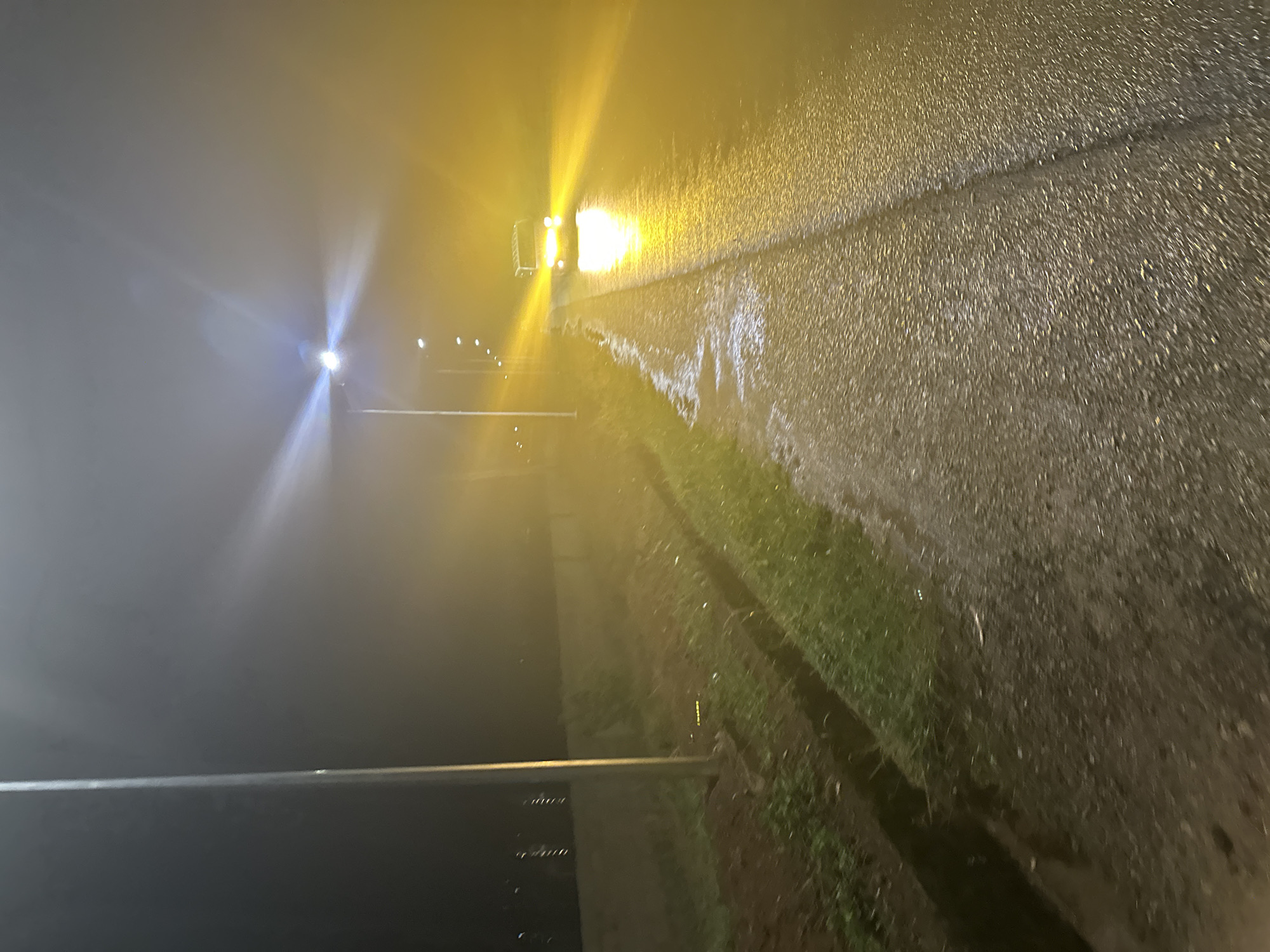 A nighttime scene, likely in Bukuru, Nigeria, presents a road surface illuminated by a vehicle's headlights. The road appears wet. Sparse grass is visible along the roadside. Streetlights are dimly visible in the distance. The overall visibility is low, possibly due to fog or rain. The image is blurry, and details are indistinct.