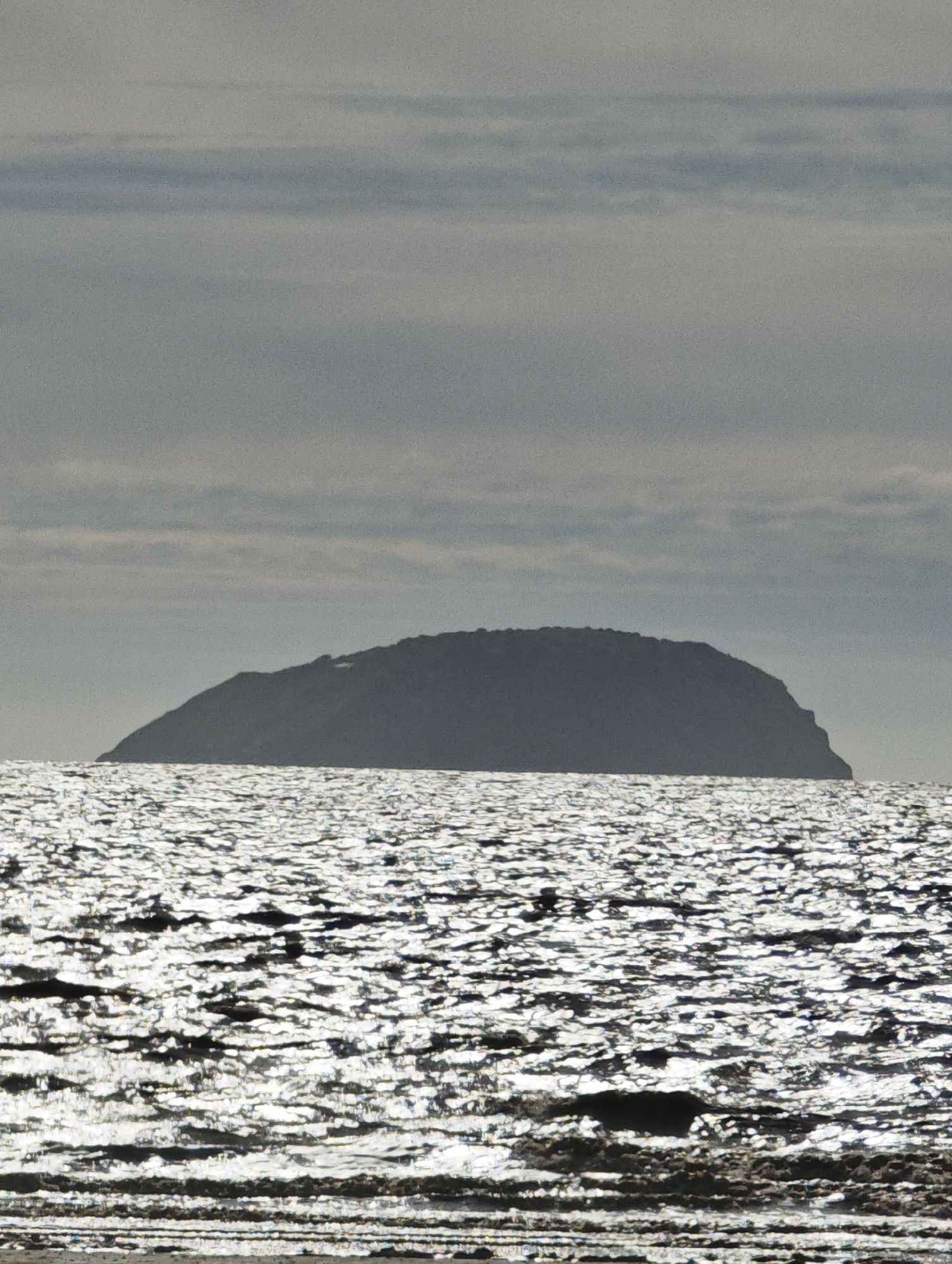 The image captures a view of the island of Steep Holm from Weston-super-Mare, United Kingdom. The island is silhouetted against a cloudy sky, the grey light adding to the solemn mood. The water in the foreground is covered in sparkling patterns due to sunlight. The foreground appears to be part of the beach, with exposed sand.