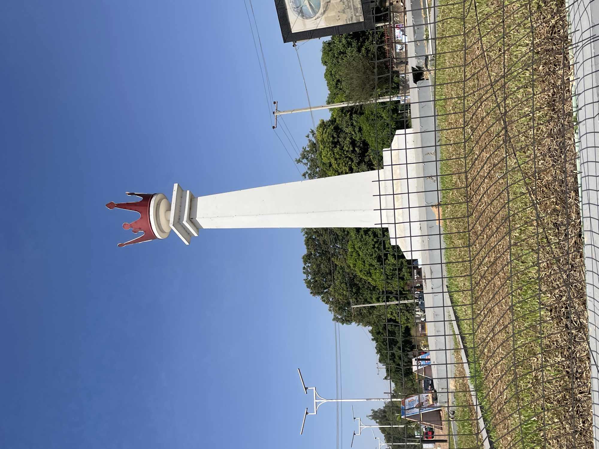 The image shows a white, elongated monument topped with a red crown-like structure. It appears to be located in Jos, Nigeria, possibly the ‘Welcome to Jos’ landmark. The monument is adjacent to a road with vehicles and power lines overhead. A black metal fence runs along the edge of the monument's area, separating it from the road. Beyond the fence, there is grass. The sky is clear blue.
