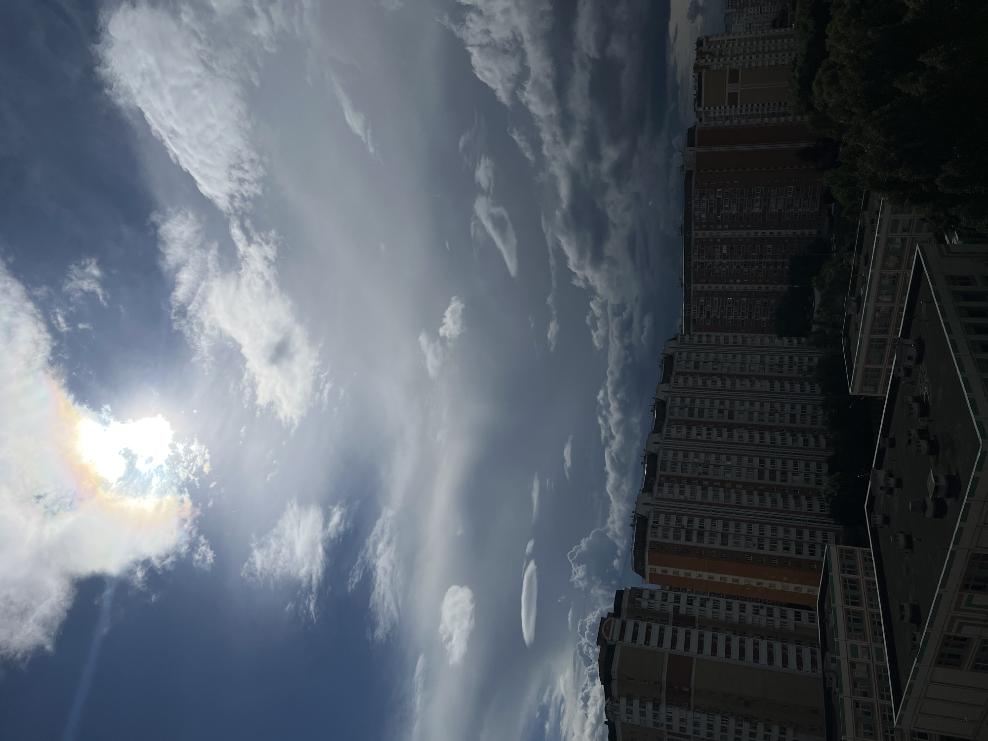 The image shows a view of a residential area in Moscow, Russia, with a dramatic sky overhead. Several tall, multi-story apartment buildings, characteristic of Soviet-era architecture, are visible in the middle ground and background. These buildings are mostly brick or concrete, with many windows. The foreground shows the flat roofs of lower buildings, possibly garages or smaller apartment complexes, with various rooftop structures such as ventilation units. Lush green trees line the horizon behind the apartment buildings. The sky is a significant feature, with a mix of dark, stormy clouds and brighter, sunlit areas. The sun is partially obscured by clouds, creating a halo effect with iridescent colors, suggesting a sun dog or similar atmospheric phenomenon. The overall impression is one of urban development under a dynamic and somewhat turbulent sky.