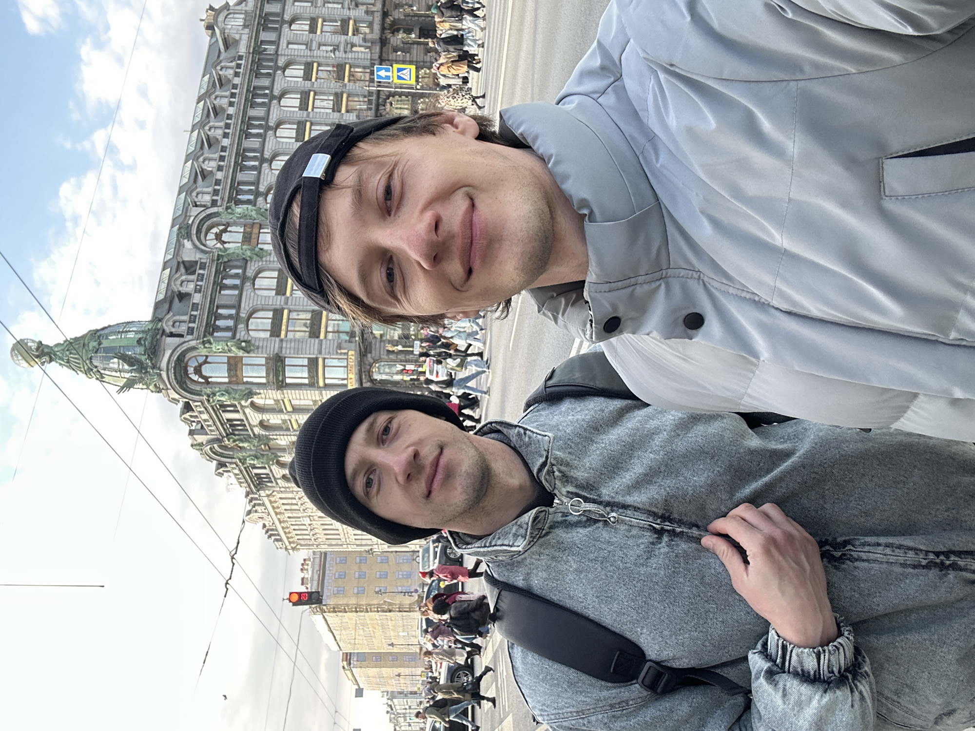 Two young men are posing for a selfie. One wears a black baseball cap and a light gray jacket, while the other wears a black beanie and a gray denim jacket with a black shoulder strap. Both are smiling. In the background is the Singer House, an Art Nouveau building, located on Nevsky Prospekt in Saint Petersburg, Russia. Pedestrians are crossing the street. A yellow pedestrian crossing sign is visible above the street. A red traffic light is visible on the left side of the frame. The sky is light blue with some clouds.