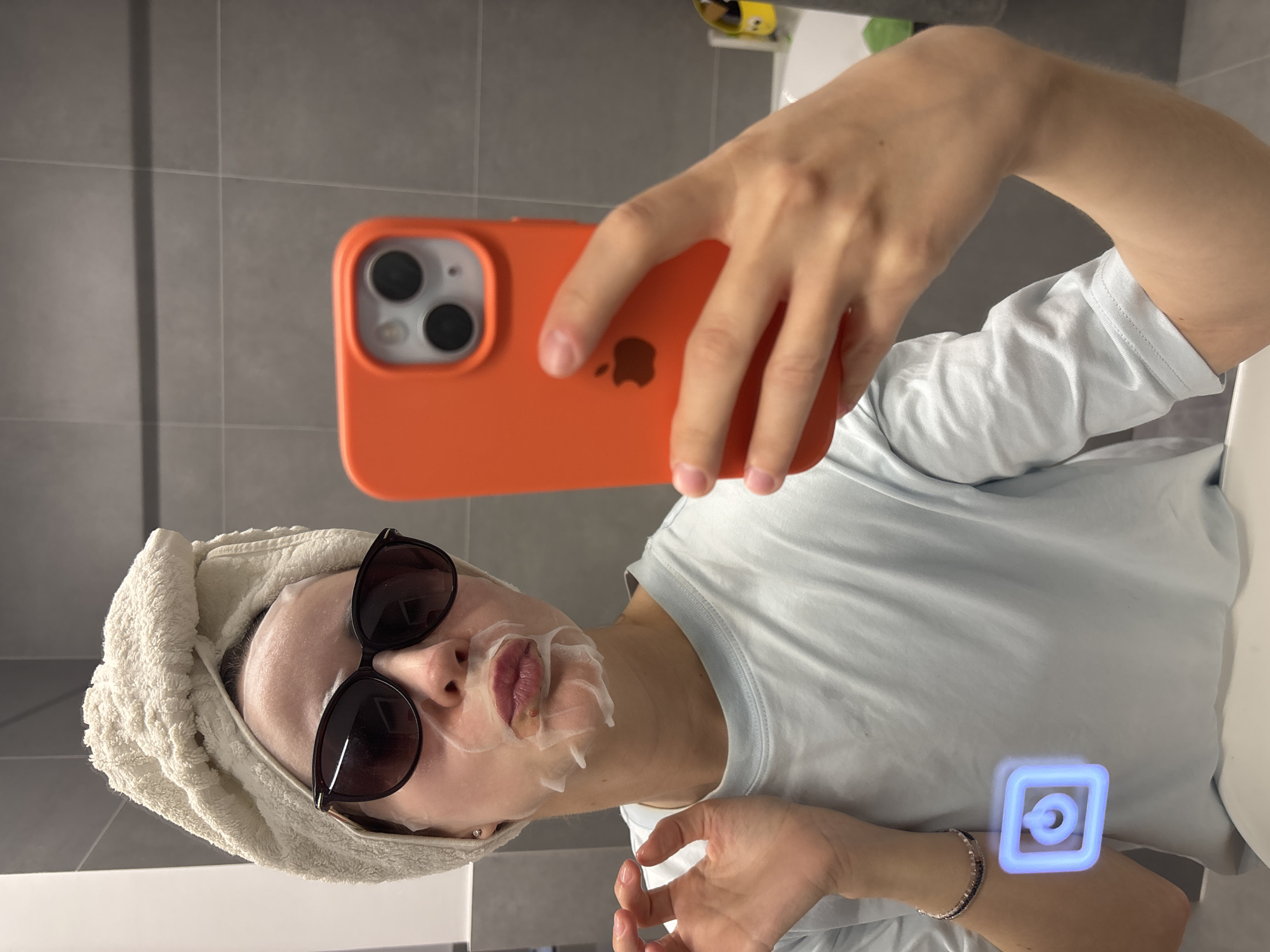 The image captures a selfie taken by a person, likely female, in what appears to be a bathroom in Chaikovsky, Russia. The individual, with fair skin, is wearing a light blue t-shirt. Her hair is neatly wrapped in a cream-colored towel. Dark sunglasses are worn over a white sheet face mask that covers most of her face, leaving her lips visible through an opening. Some residue from the mask is visible around the mouth area.
The person is holding an orange-cased iPhone in her right hand to take the selfie. The distinct Apple logo is visible on the back of the phone case. The background features a grey tiled wall and a white surface, possibly a sink or countertop. On the lower right side of the image, a glowing blue icon resembling a social media logo or a power symbol is digitally superimposed. A multi-colored beaded bracelet is visible on the person's left wrist. The scene suggests a moment of self-care or relaxation. No specific time of day or weather details are discernible.