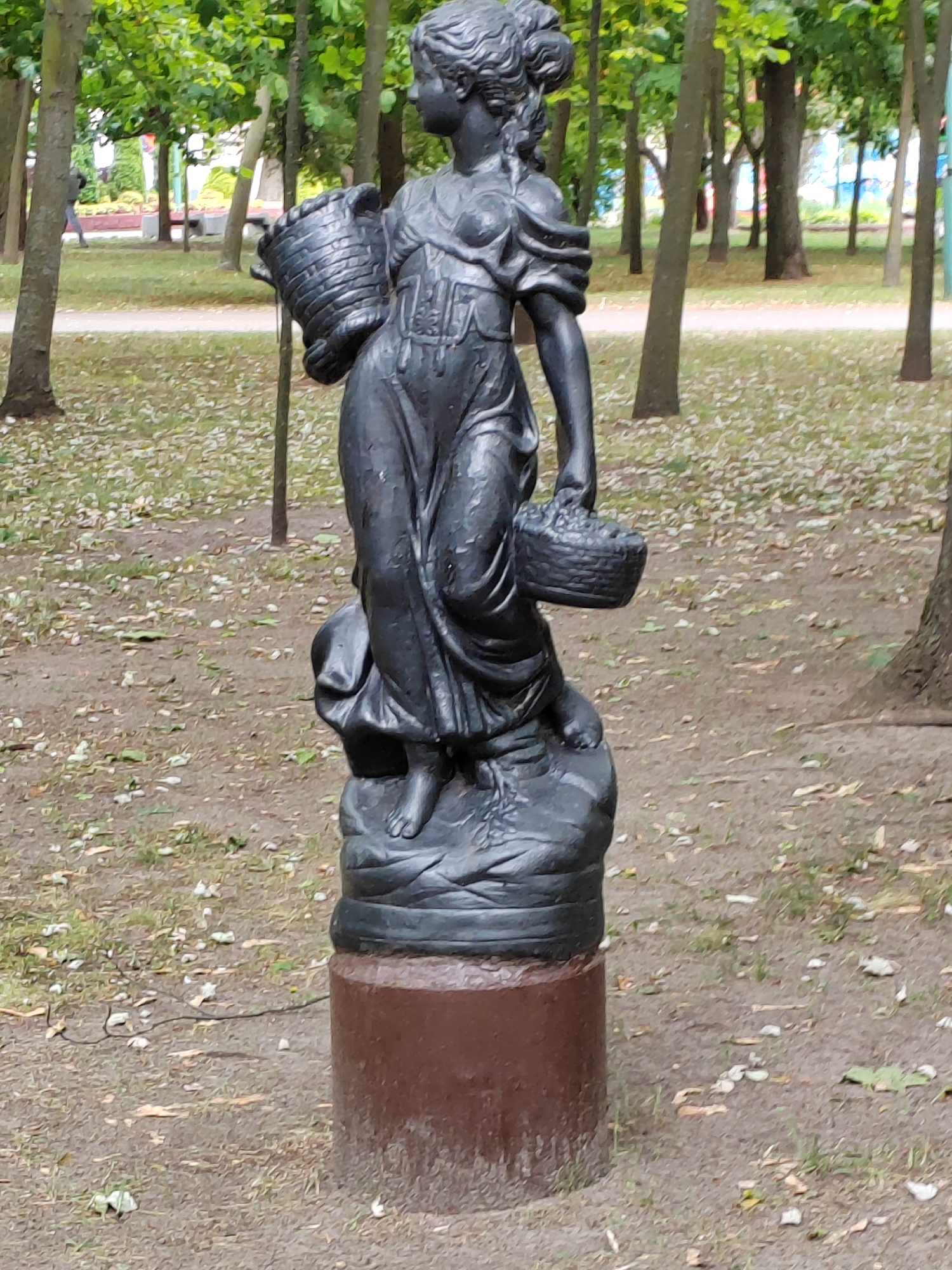 The image shows a dark gray, weathered statue of a young woman standing on a pedestal in an outdoor park setting. The statue depicts the woman wearing a long, flowing dress and holding a woven basket in her right arm and another basket in her left hand. Her hair is styled in an updo. She stands on a base that appears to be a dark, rocky formation. The pedestal is a cylindrical object, reddish-brown in color, with some visible wear and tear. The background of the image consists of a park with trees, grass, and fallen leaves on the ground. The trees are slender and appear to be deciduous. Sunlight filters through the leaves, casting shadows. In the distance, there are hints of man-made structures, possibly buildings or park amenities, and a walking path. The overall scene suggests a public park in Brest, Belarus, likely a historical or decorative element within the city's green spaces.