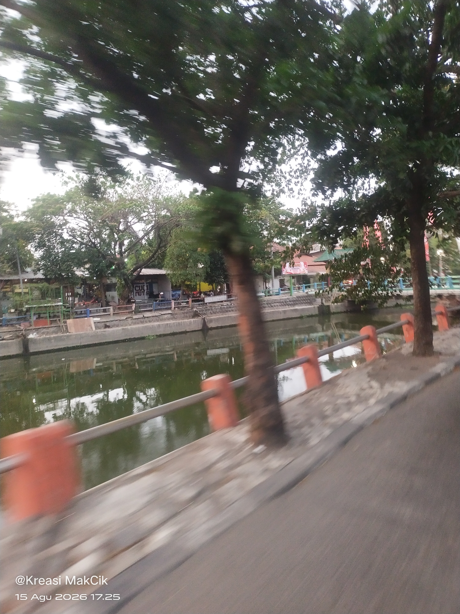 The image depicts a waterfront scene, likely in Surabaya, Indonesia, given the timestamp "@Kreasi MakCik 15 Agu 2026 17.25". The water appears somewhat murky and reflects the surrounding trees and structures. A concrete embankment lines the waterway, and a railing with orange posts is visible. Beyond the railing, there are trees, some with dense foliage, and buildings that appear to be small shops or stalls. The image has a motion blur, suggesting it was taken from a moving vehicle. The overall impression is of a bustling, possibly commercial, area along a canal or river.