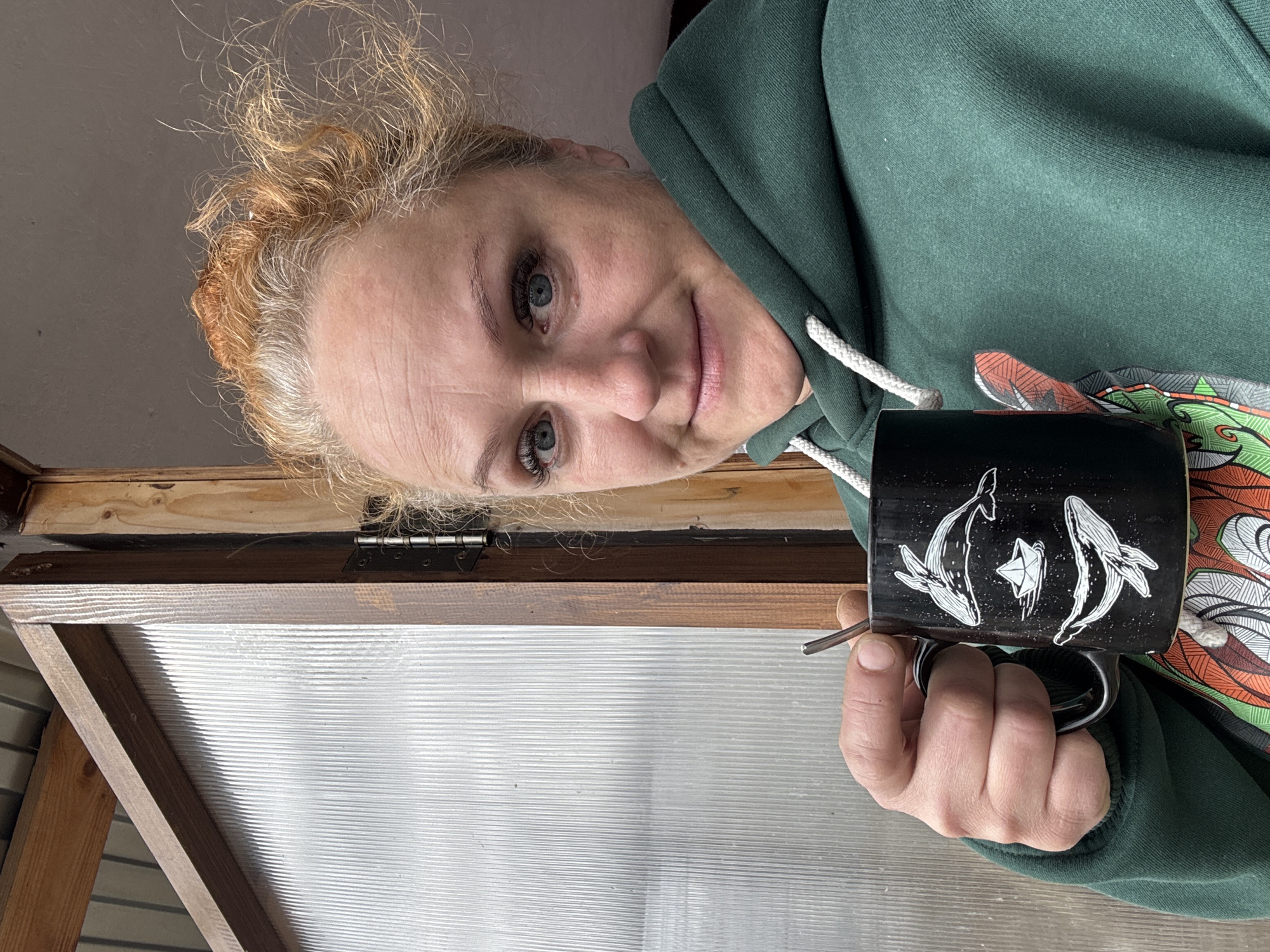 A woman with light skin, light eyes, and curly blonde-reddish hair is holding a black mug with white whale designs and a small spoon. She is wearing a green hooded sweatshirt with white drawstrings and what appears to be a colorful design peeking from underneath. The background shows a window-like structure with a wooden frame and a corrugated plastic or similar material as the pane. The scene is likely indoors, possibly a greenhouse or a shed-like structure. The woman is looking at the camera with a slight smile. The location is Chaikovsky, Russia.