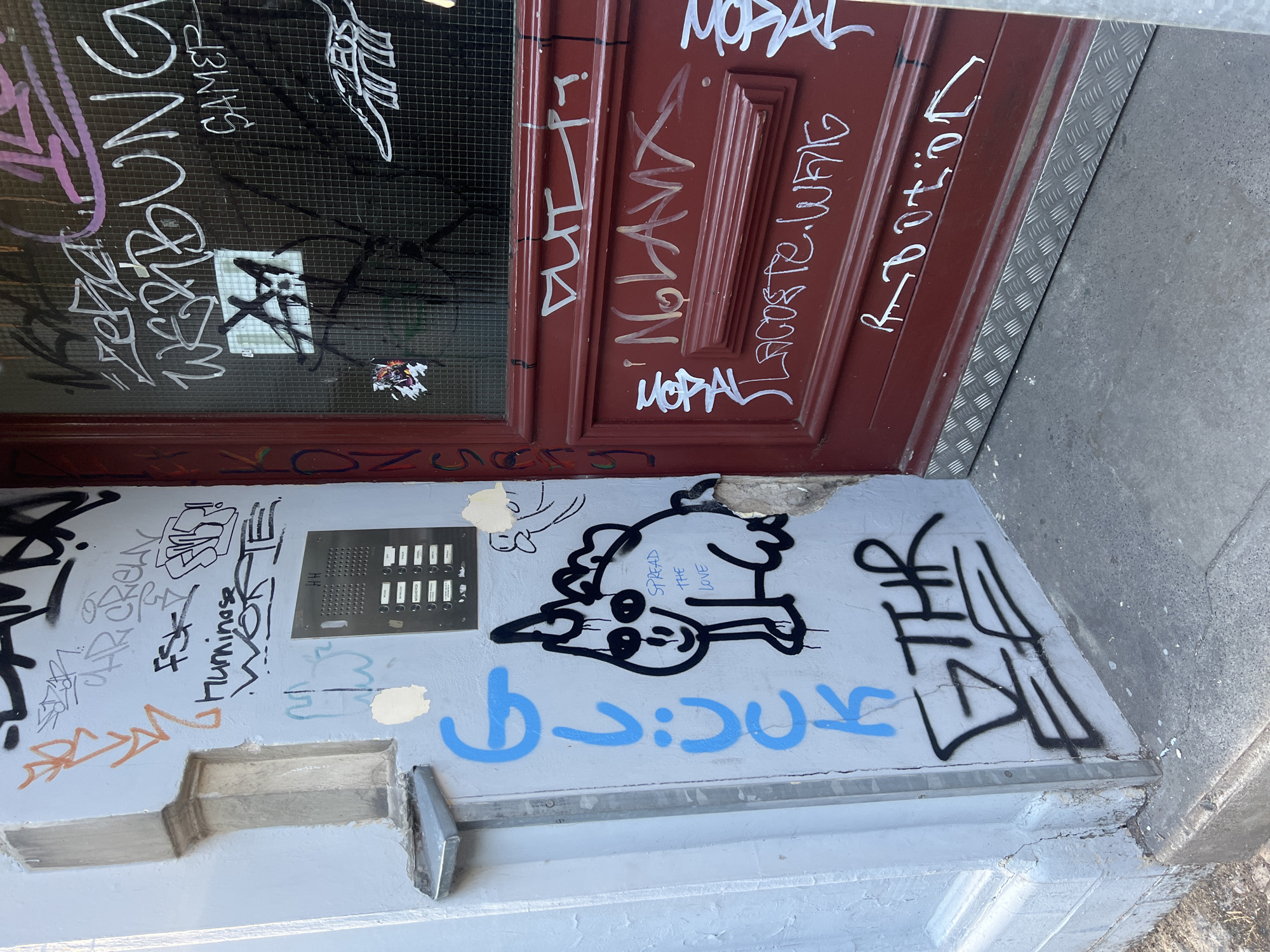 The image shows the exterior of a building entrance in Berlin, Germany, characterized by significant graffiti on the wall and the wooden door. The wall to the left is a light blue-grey, with a small, weathered metal downspout fixture and architectural molding. The door is a dark, rich red.
Dominating the scene is an abundance of graffiti in various colors including black, white, purple, and orange. On the blue-grey wall, a stylized blue word "GLÜCK" is written vertically, accompanied by a black and white cartoonish drawing of a creature with a unicorn horn and the text "SPREAD THE LOVE" written inside it. Below this, in black graffiti, is the word "ETHR".
The red door is heavily tagged with white and light-colored graffiti. Visible tags include "WERBUNG SAMER", "xzfa", "X", "ZEUS", "NOLAVX" with a heart shape, "LACOSTE.WUIG", and "R.OBOTion". There are also smaller stickers and scribbles on the door. The entrance area is at street level, with concrete pavement and a metal kick plate at the bottom of the door. The overall impression is one of urban decay and artistic expression through graffiti.