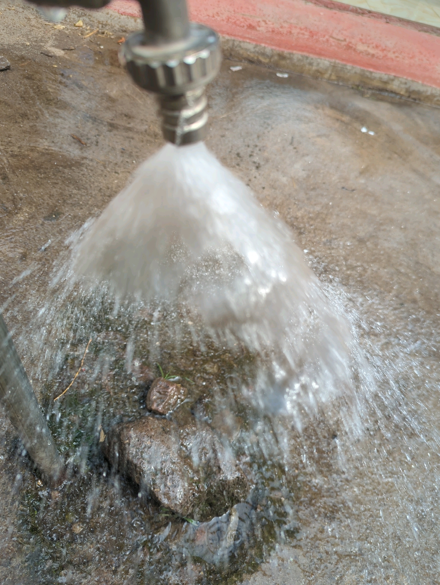 A close-up, overhead shot shows a metal faucet spewing a wide stream of water onto a concrete surface. The water creates a mist and splashes as it hits the ground, which appears to be gritty and uneven, with small pebbles and some sparse vegetation visible. In the background, a red border or edge is visible, suggesting the presence of a raised planter or walkway. The overall scene is wet and slightly chaotic due to the force of the water. The location is Musanze District, Rwanda.