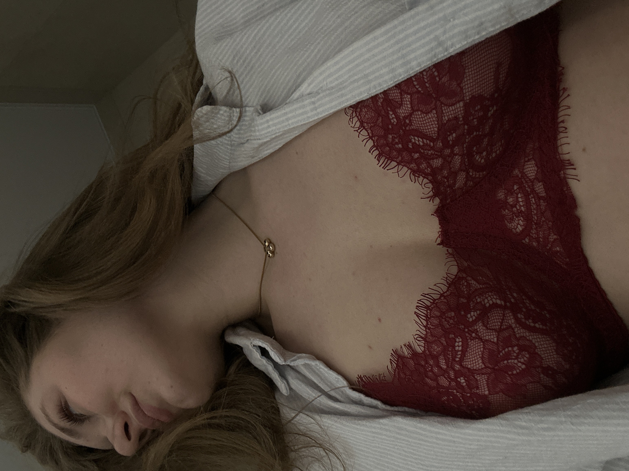 A woman with long, light brown hair is lying down, possibly in bed, wearing a striped collared shirt that is partially unbuttoned, revealing a red lace bra. A gold necklace is visible around her neck. The background is a plain wall. No information in the image suggests the location is Kazan, Russia.
