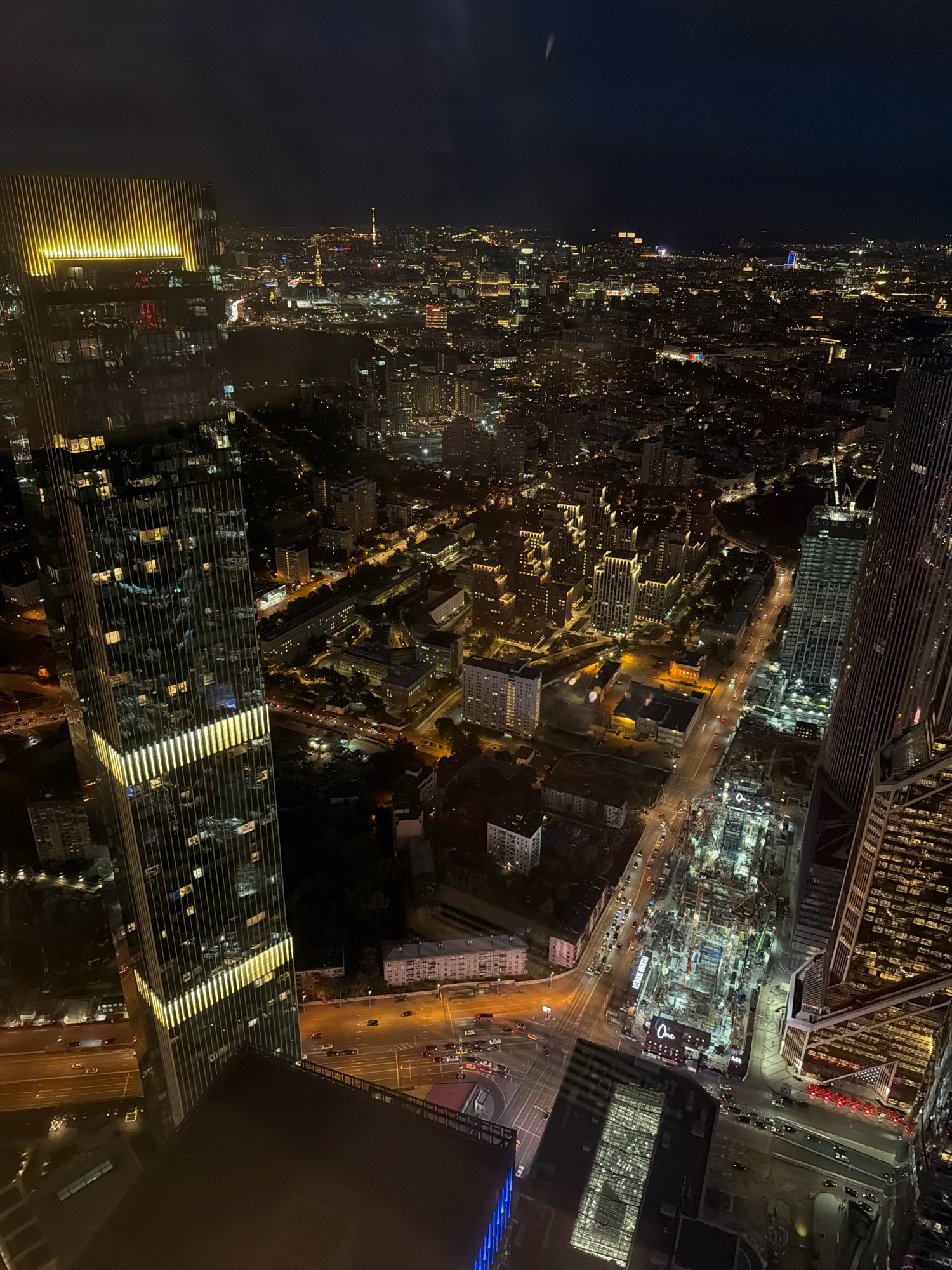 This aerial photograph captures a night view of Moscow City, Russia, with a prominent skyscraper in the foreground and a sprawling urban landscape extending into the distance. The skyscraper is modern, with a glass facade reflecting the city lights and vertical lighting strips. Below, the city is illuminated by countless streetlights and building lights, showcasing a dense network of roads, buildings of various heights, and what appears to be a construction site with cranes. The dark sky suggests it is nighttime, with a few distant lights visible on the horizon. The perspective is from a high vantage point, looking down on the illuminated urban expanse.