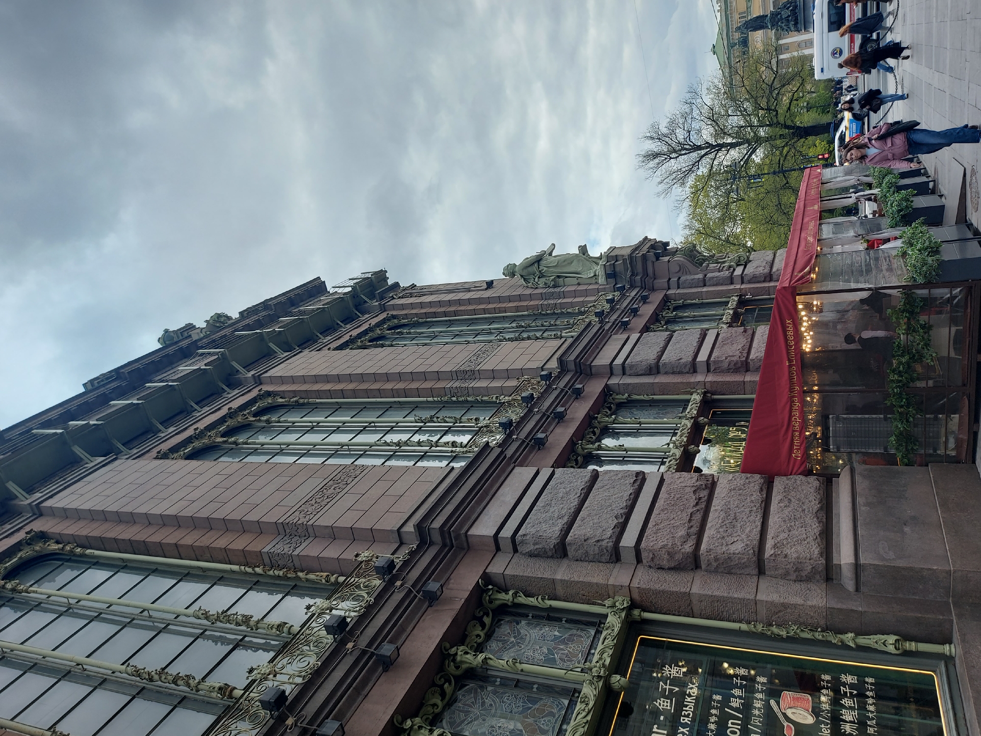This image depicts the Eliseev Emporium in Saint Petersburg, Russia. The building is shown from a low angle, highlighting its ornate facade with rows of large arched windows, elaborate metalwork, and stone detailing. A statue is visible on the roofline. On the right side of the image, there is a section of the building's exterior that extends outwards, with a red awning and glass enclosures, likely a restaurant or cafe space. The reflection on the glass contains lights and decoration.
On the sidewalk, people are walking by. A few cars are also visible in the background behind a large tree. The sky above is overcast with grey clouds. The signage on the glass walls includes text in both Russian and Chinese, advertising seafood and caviar.