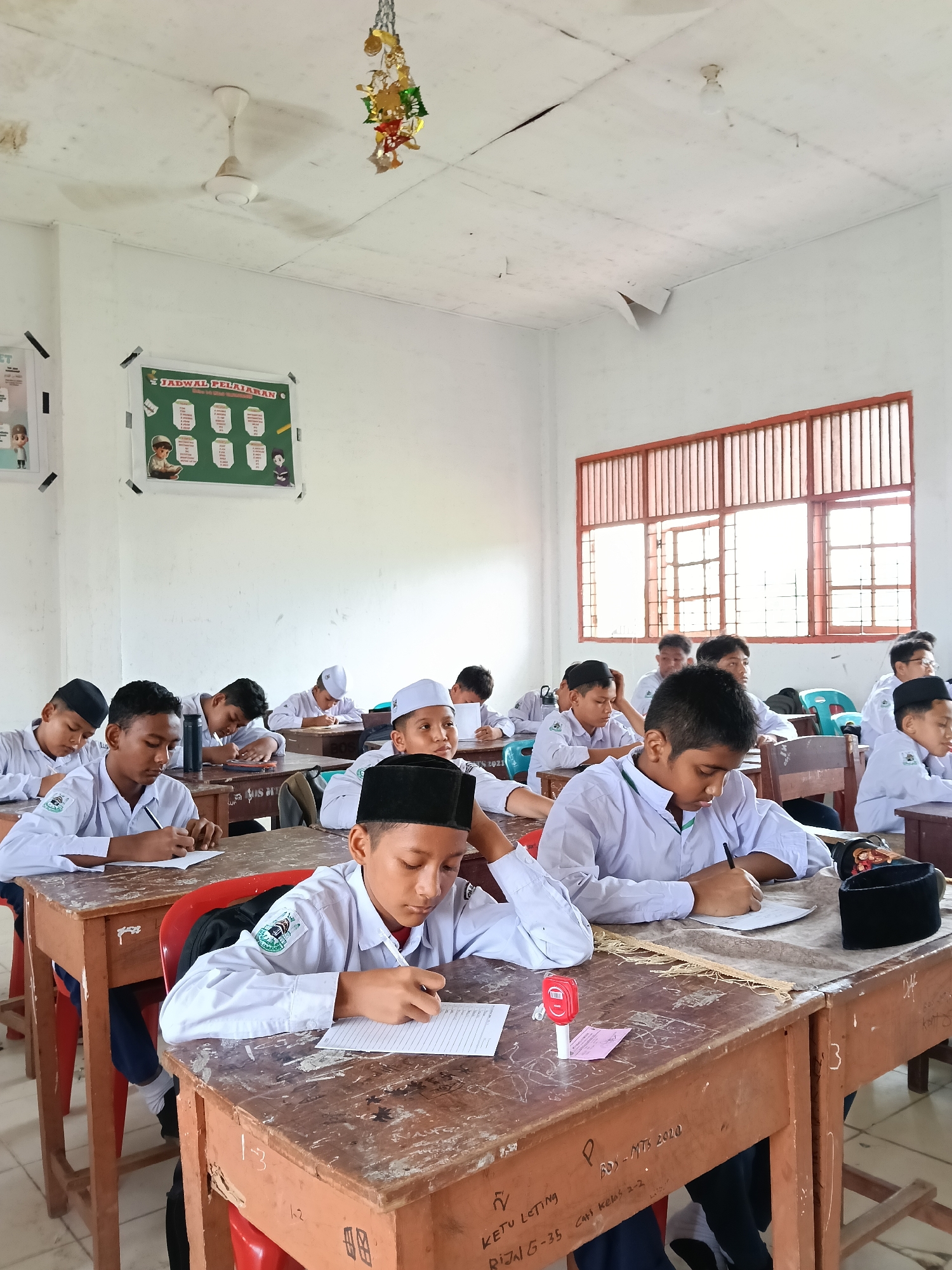 The image depicts a classroom scene in what appears to be a school in Lhokseumawe, Indonesia. Several young male students, presumably in their early teens, are seated at wooden desks and engaged in writing. Most are wearing white shirts, and some have head coverings, indicating a religious or cultural association, and likely indicating their enrollment in a pesantren (Islamic boarding school). The classroom's walls are white, with a timetable poster displayed. The desks show signs of wear, including writing and markings. Outside the window, there is natural light suggesting it is daytime.