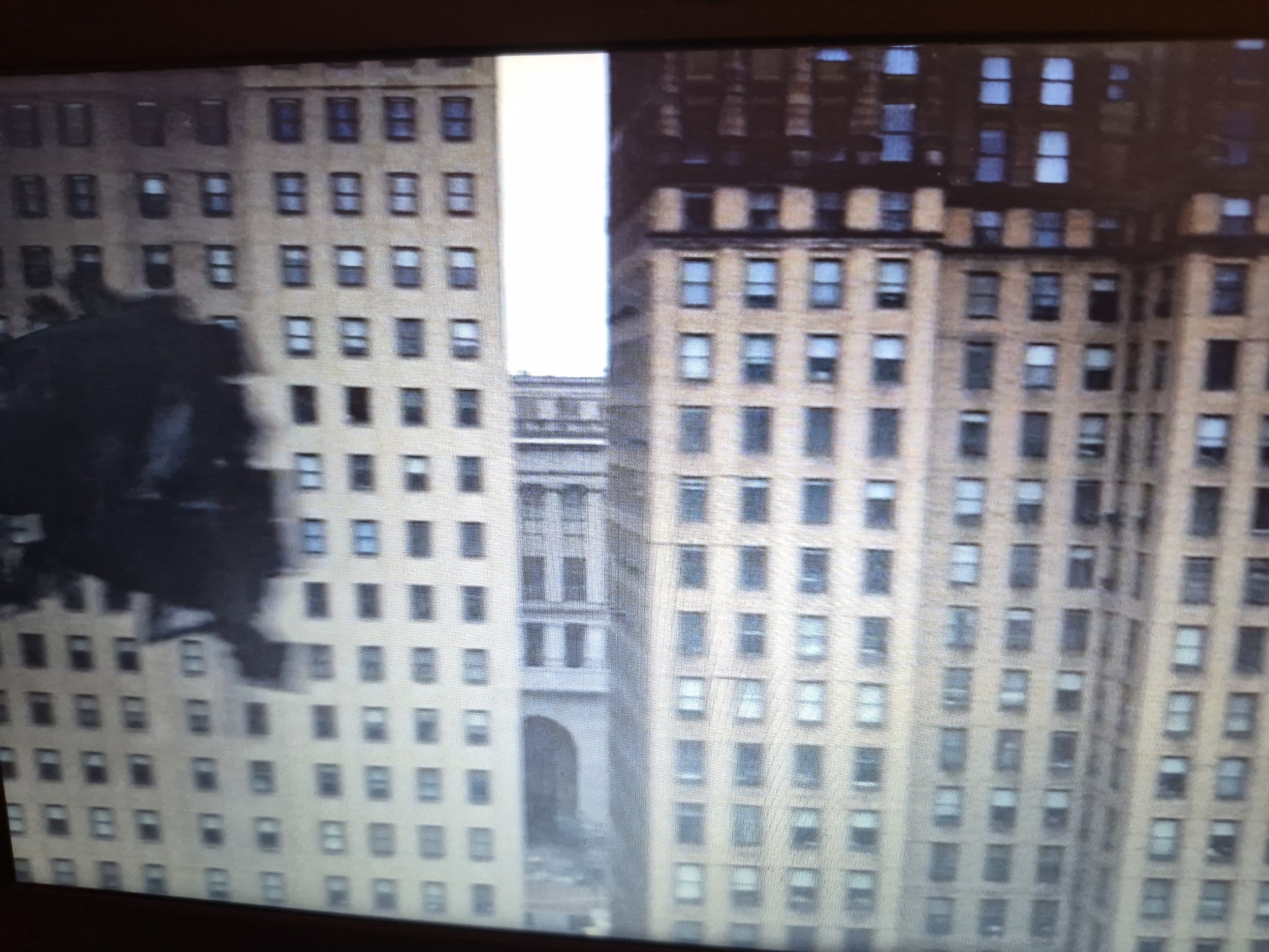 The image shows a close-up, slightly blurry, aerial view of tall, beige brick buildings with many windows. The buildings appear to be in a dense urban environment. On the left side of the image, a dark, amorphous shadow or object is superimposed on one of the buildings, partially obscuring its windows. The buildings are arranged in rows and columns of rectangular windows, with some variation in the window shapes and sizes. The building on the right has a darker brick facade and a more ornate architectural style at the top. A narrow, brightly lit vertical gap between the two main buildings reveals a glimpse of another structure with an arched entryway. The context of Sandwell, United Kingdom, is not visually evident in this image.