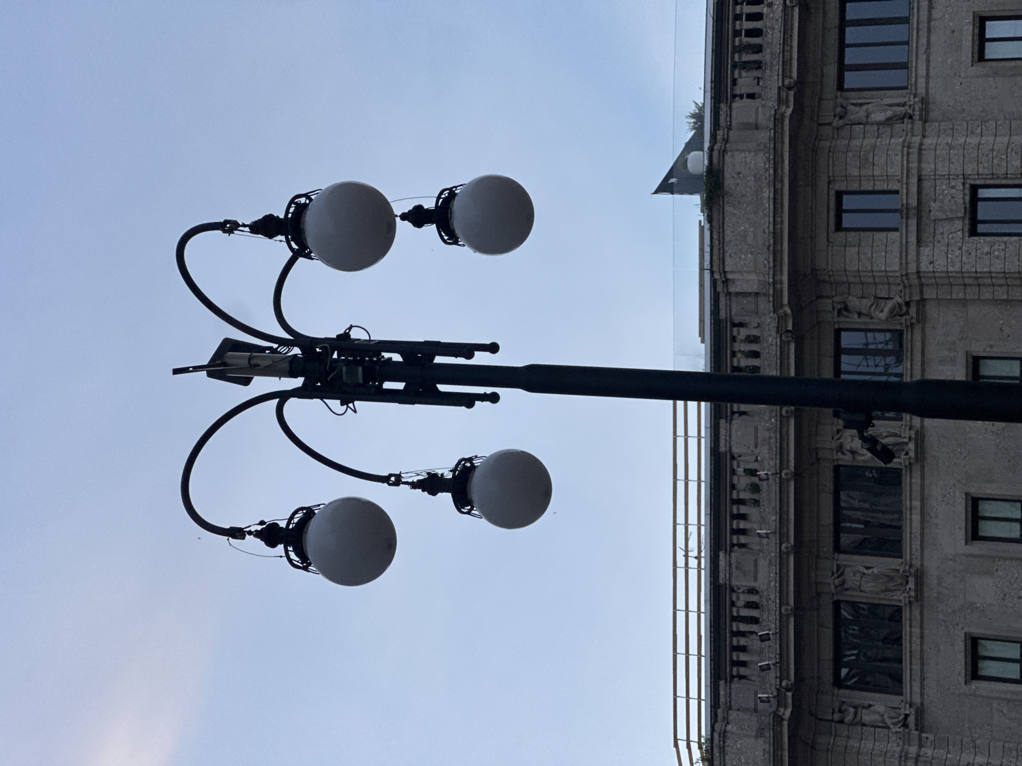 A street lamp with four spherical white lights stands tall against a pale blue sky, its black metal frame curved to support each lamp. In the background, a classical-style building with ornate stonework, arched windows, and statues is visible. The building appears to be an old, well-maintained structure, possibly a historical landmark. The overall scene is serene and captures a moment of stillness in an urban environment. Based on the architectural style and the presence of street lamps of this design, this scene is likely in Milan, Italy.