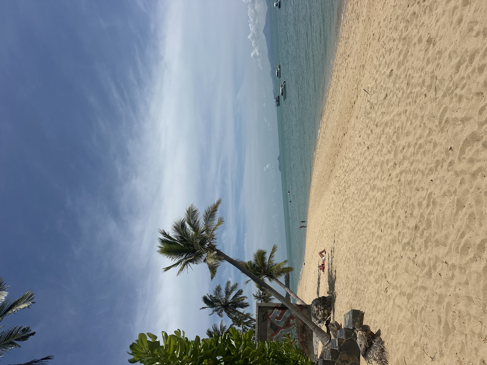 The image depicts a sandy beach with palm trees in the foreground. The ocean stretches out to the horizon, with several boats visible on the water. In the distance, mountains can be seen. The sky is blue with scattered clouds. There are a few people on the beach, some of whom are swimming. The overall scene is one of a tropical paradise. This is a beach in the Ko Samui District of Thailand.