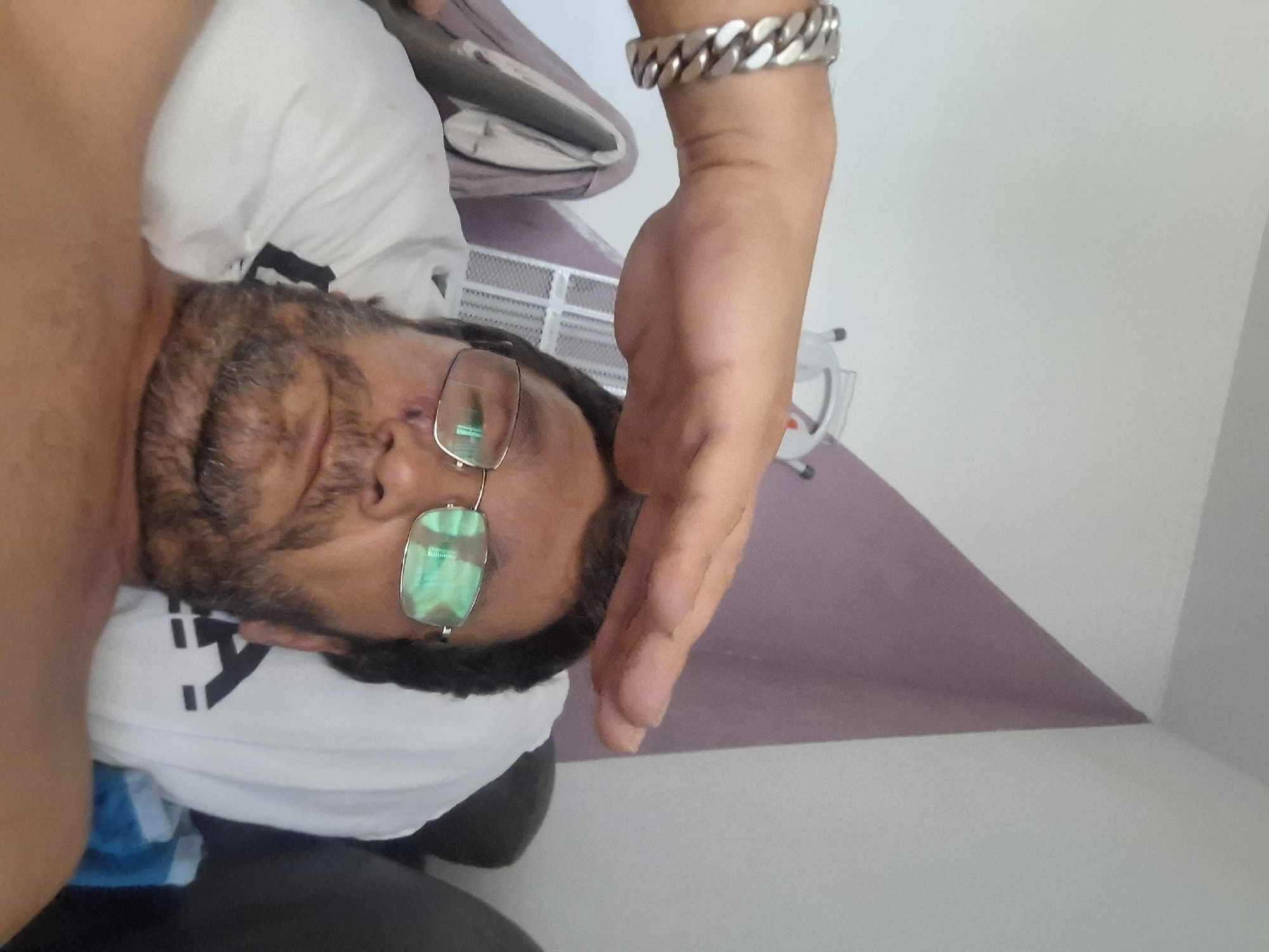 A first-person perspective selfie shows a man with a beard and glasses lying down in what appears to be a bedroom. A hand with a silver bracelet is placed on top of his head, as if to comfort or bless him. The man has a band-aid on his cheek and a bruise near his eye. The background features a wall with a purple accent and a white ceiling. The scene is consistent with a domestic setting in Yerocham, Israel.
