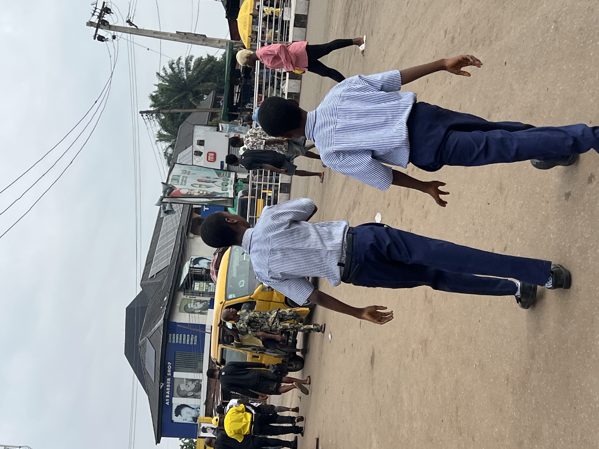 In Nkpor, Nigeria, on a cloudy day, two young boys wearing blue and white striped shirts and navy blue pants walk away from the camera down a dusty, unpaved street. The boy on the left has his left arm bent at the elbow and his right arm extended slightly out to his side. The boy on the right has both arms extended slightly out to his sides, with his left hand dangling down and his right hand held up. Several other people are visible in the background. To the left, a yellow van is parked on the side of the road, with people entering and exiting it. A man in a camouflage uniform is standing next to the van. Further back, a building with blue siding and a dark-colored roof has an "A1 BARBER SHOP" sign on it, with images of hairstyles displayed. Other businesses are visible beyond the barber shop, including one with a large billboard featuring faces. Power lines and utility poles crisscross the sky above the scene. A yellow umbrella is visible in the far right of the image.