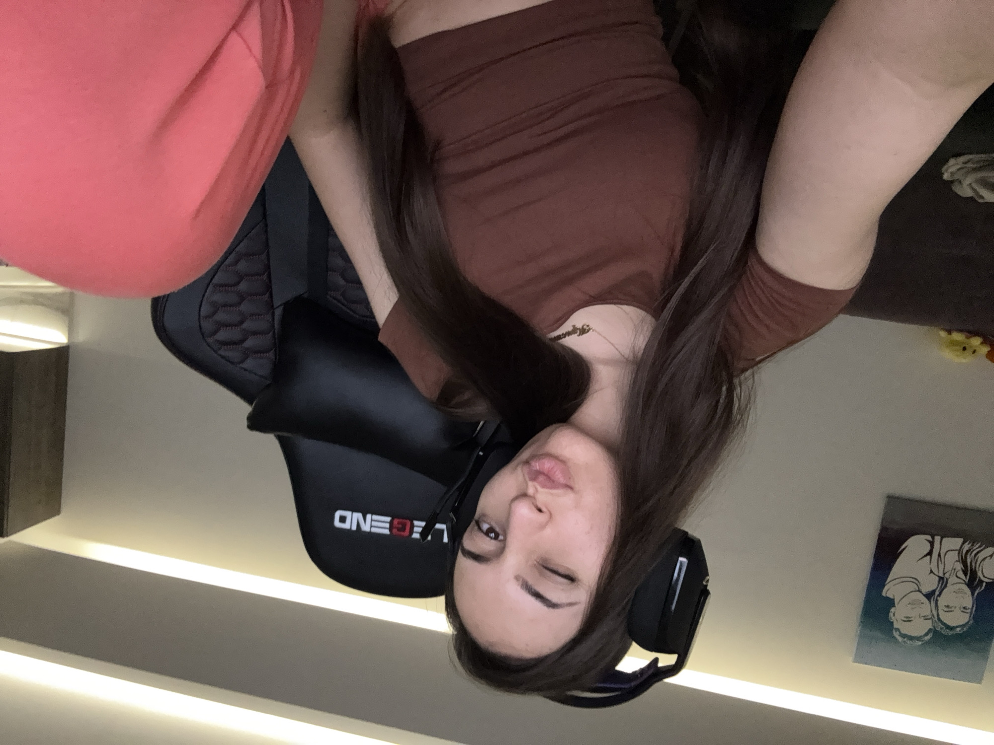 A young woman with long brown hair is sitting in a black gaming chair with the word "LEGEND" printed on it. She is wearing a brown cropped top and pink pants. She is winking and wearing headphones. Behind her is a wall with a painting on it. The image appears to be taken indoors, under an artificial light. There is no verifiable information in the image to confirm the location in the city of Plovdiv, Bulgaria.