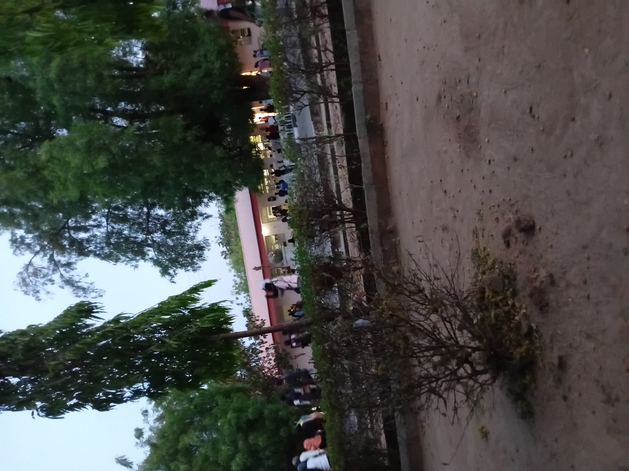 A ground-level view shows a dirt path on the right and a row of bushes separating the path from what appears to be a building with people gathered outside. The sky is overcast, and trees are visible on the left of the image. The building has a red roof and lights are on inside. Some people are walking or standing, while others seem to be seated or gathered in groups.