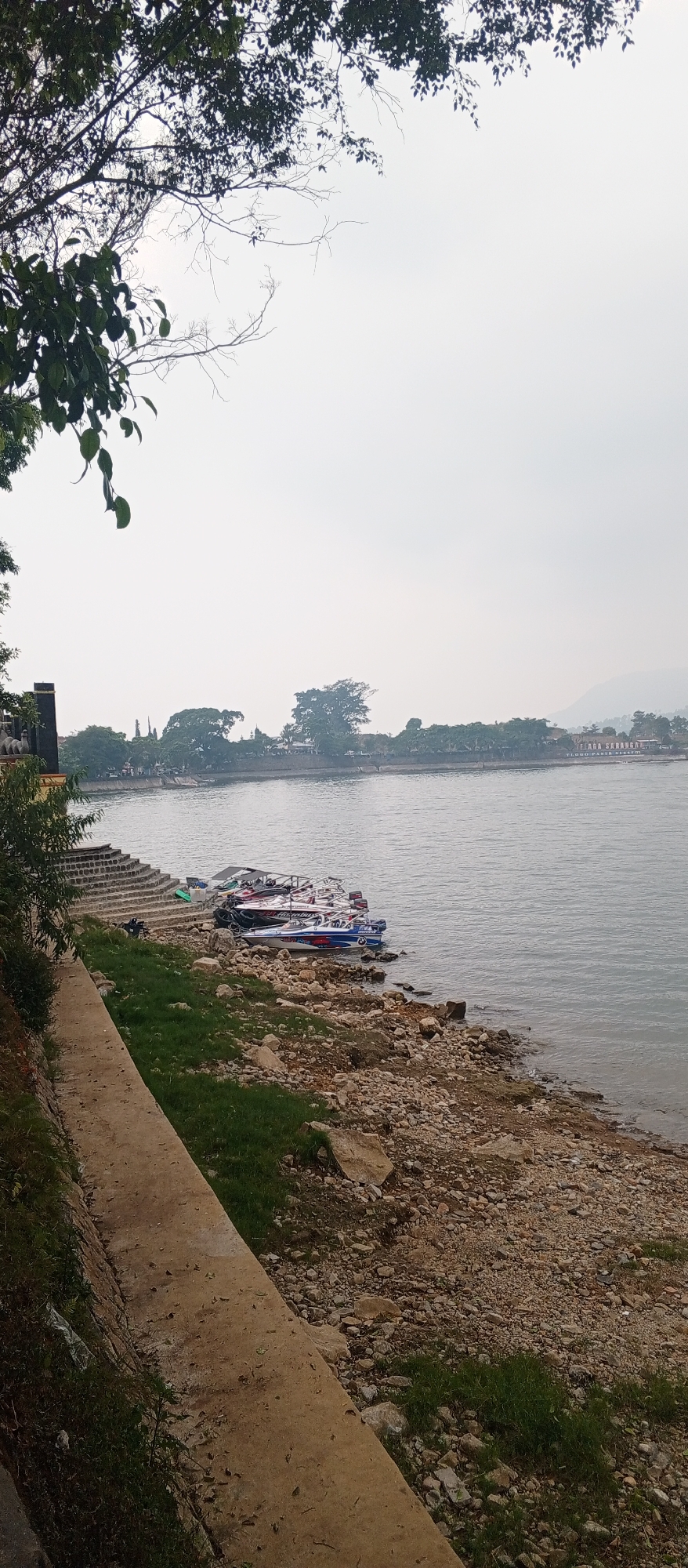 The image depicts a waterfront scene in Sarangan, Indonesia, a popular tourist destination known for its lake. The foreground features a concrete walkway alongside a rocky, grassy shoreline. Several motorboats are moored along the edge of the water, some appearing to be speedboats. In the middle ground, a calm body of water extends towards the opposite bank, which is lined with trees and buildings. The background is dominated by a hazy sky and distant hills. The overall atmosphere is serene, with no people visible in the frame.