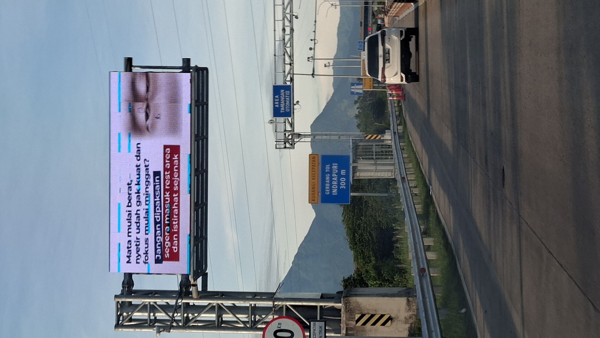 The image shows a digital billboard on a highway with a message about driver fatigue. The billboard displays text in Indonesian that translates to "Eyes starting to feel heavy, already can't stand driving and focus is starting to waver? Don't force it, immediately enter a rest area and rest for a while." The background of the billboard features a close-up image of a person's tired eyes.
Below the billboard, there are road signs indicating "KURANGI KECEPATAN" (Reduce Speed) and "GERBANG TOL INDRAPURI 300 m" (Indrapuri Toll Gate 300 m). There is also a sign for "AREA TIMBANGAN OTOMATIS" (Automatic Weighing Area).
The highway itself is visible, with a white SUV driving on the right lane. The environment appears to be mountainous with some vegetation. The sky is clear and bright, suggesting it's daytime. The location is identified as Sihomlhok, Indonesia.