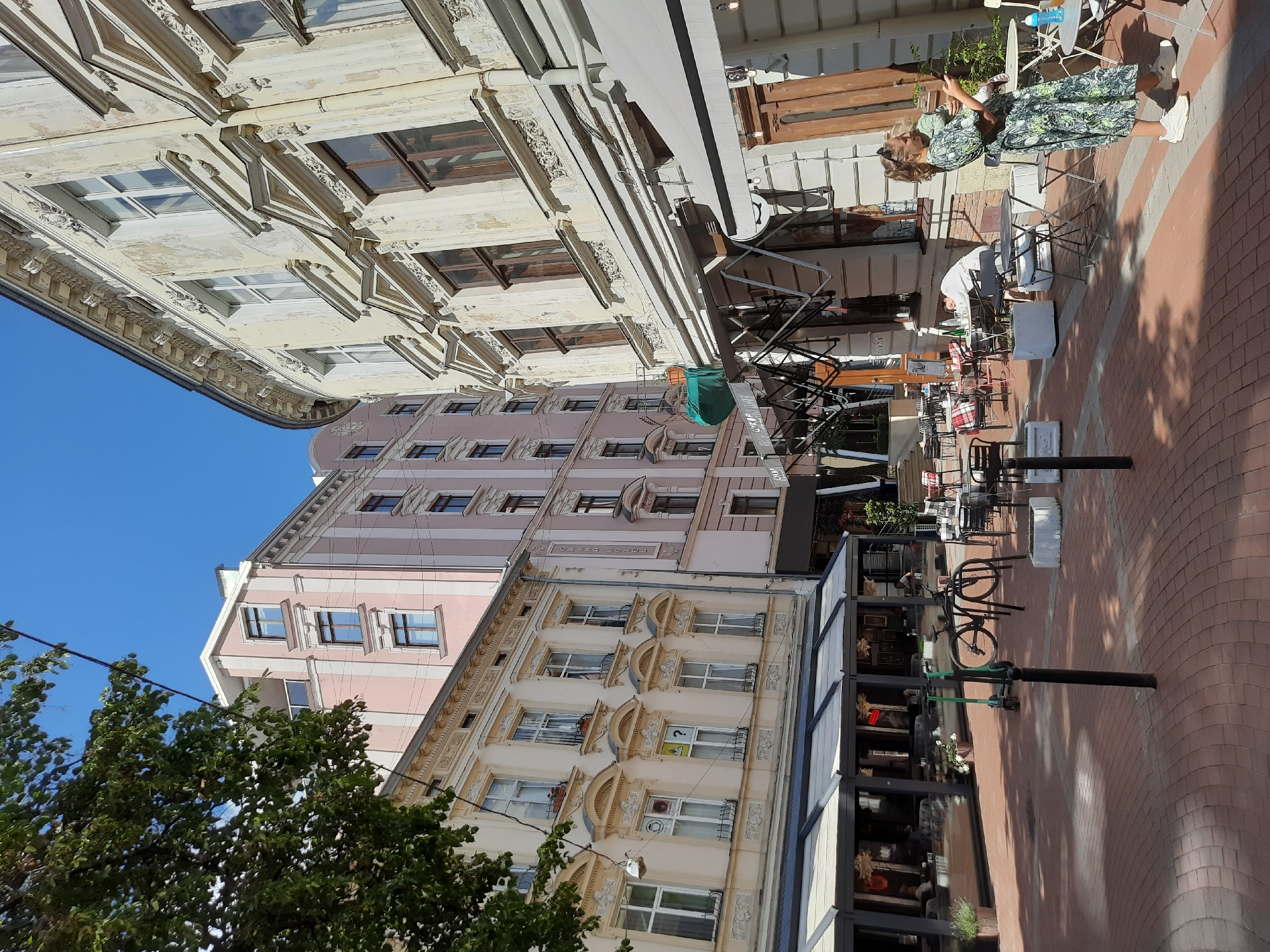 The image captures a sun-drenched scene on a cobblestone street in Lviv, Ukraine, likely a pedestrian zone with outdoor seating for cafes. In the foreground, a woman in a patterned dress stands holding a baby, looking at her phone. She is wearing white sneakers. To her left, several outdoor cafe tables and chairs are visible, with some patrons seated. Further back, a bicycle and an electric scooter are parked. The street is lined with historic buildings, some displaying signs of age with faded paint and ornate architectural details. One building to the left is painted in pale yellow and adorned with classical moldings. Another building, further back and to the left, is a taller, modern structure painted in a light pink hue with white vertical stripes. To the right, a building with a curved facade features weathered, creamy-colored walls and dark-framed windows. The sky above is a clear, bright blue. A large green tarp is suspended from scaffolding in front of one of the buildings.