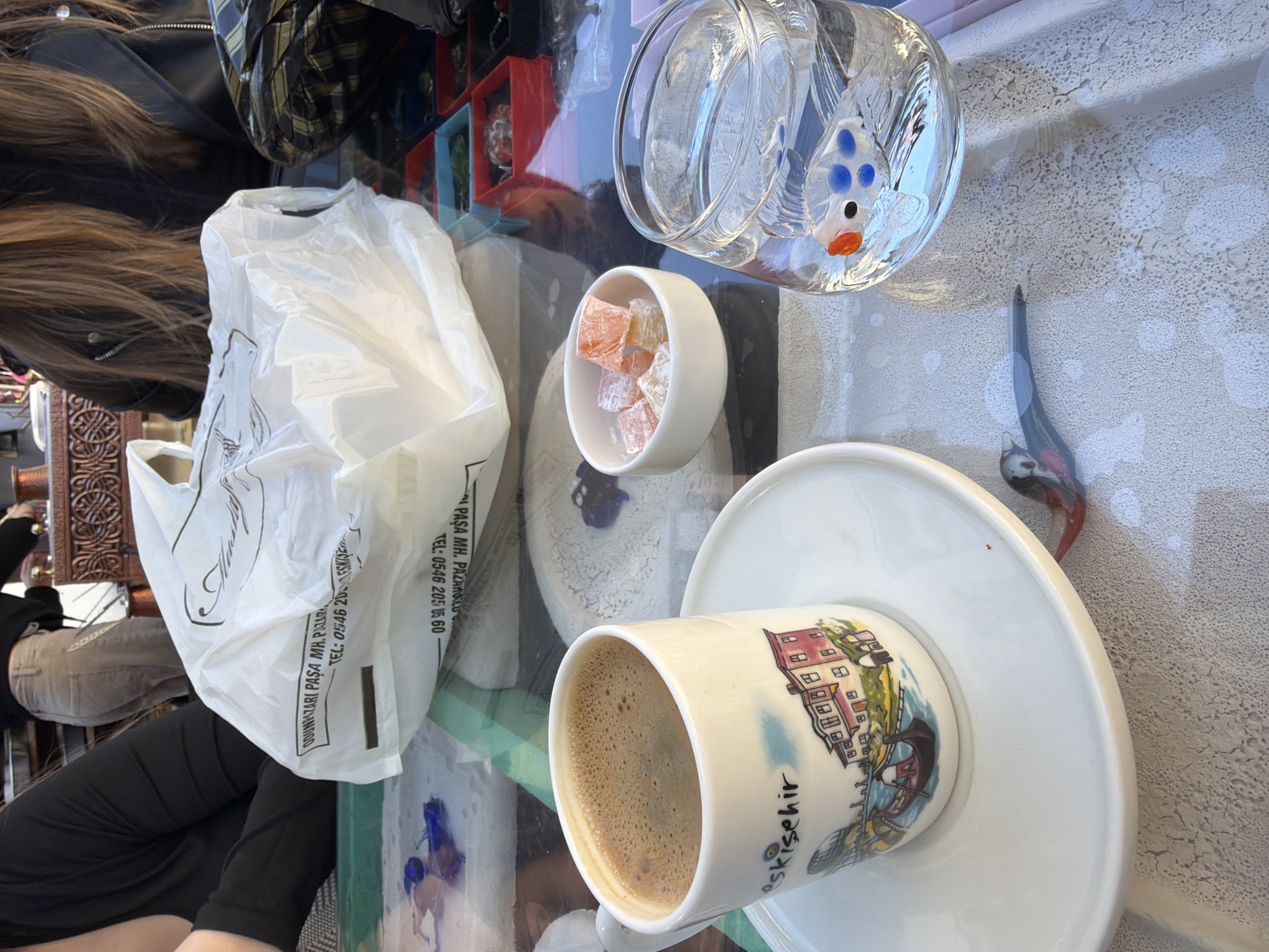The image captures a vibrant tabletop scene, likely outdoors or in a well-lit cafe, with a person partially visible in the frame. A white ceramic cup of foamy Turkish coffee rests on a matching saucer, featuring a colorful illustration of buildings and the word "Eskişehir" printed on its side, confirming the location as Eskişehir, Turkey. Adjacent to the coffee, a small white bowl holds several cubes of colorful Turkish delight (lokum) in orange and light pink hues.
To the upper right, a clear glass ornament resembling a fish, with distinctive blue and orange accents, is visible. A slender, decorative bird figurine, possibly made of ceramic or wood, also sits on the table. A white plastic shopping bag, partially crumpled, occupies a significant portion of the left side of the table. The bag displays text in Turkish, including "OĞUZARİ PAŞA MH. PAZARNALO TEL: 0546 205 05 60" and "PAŞA MH. TEL: 0546 20," which likely denote a business name, address, and phone number.
The background shows glimpses of the person's long brown hair, part of a black jacket, and a lower leg in black trousers, suggesting a casual setting. The table surface appears reflective, capturing ambient light and faint reflections of other objects. Bright, natural lighting indicates it is daytime. The overall scene suggests a leisurely moment, enjoying traditional Turkish refreshments.