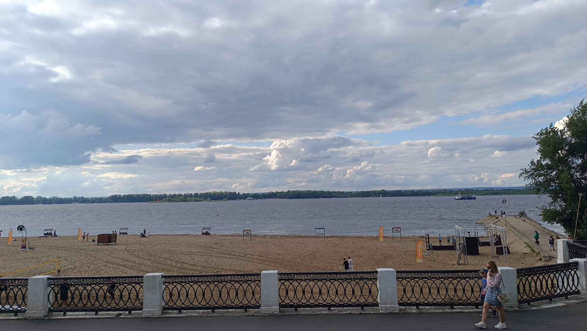 The image captures a view of the Volga River beach in Samara, Russia, on a cloudy day. The foreground features a black ornamental metal fence with white decorative pillars. Beyond the fence is a sandy beach with scattered beach umbrellas, volleyball nets, and a few people. In the middle ground, the wide Volga River stretches to the horizon, with a distant treeline on the opposite bank. A blue passenger boat is visible on the water, along with a long pier extending into the river with several people on it. The sky is filled with a mix of dark gray storm clouds and patches of lighter, brighter clouds, suggesting an overcast or partly cloudy day with potential for rain.