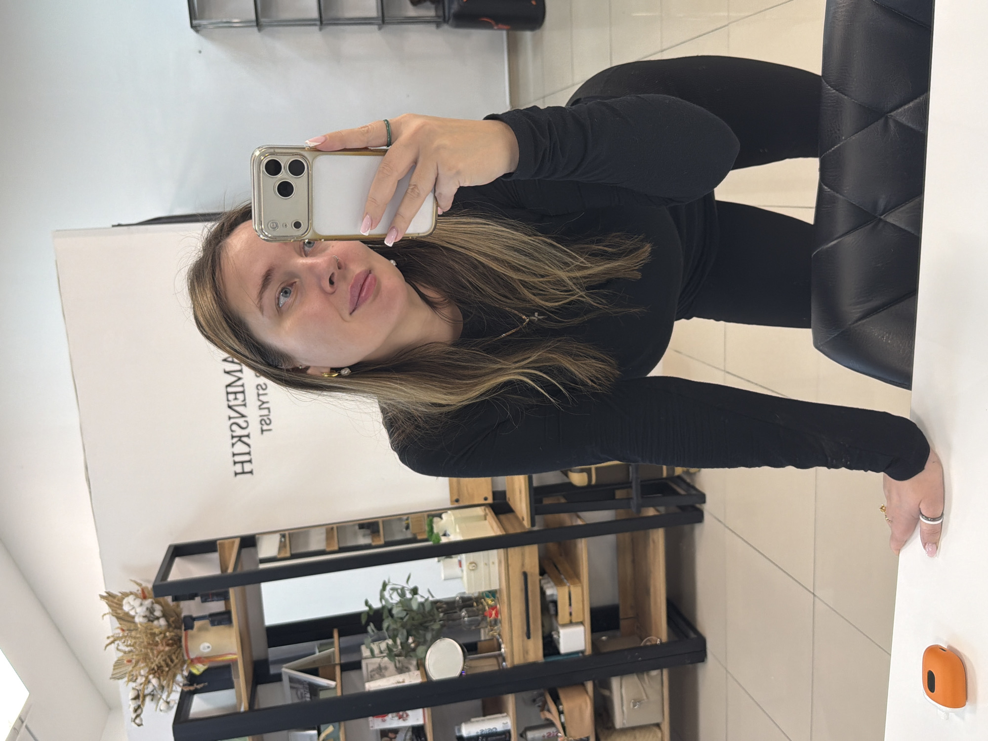 An indoor scene in Chaikovsky, Russia, depicts a woman taking a selfie in a mirror. The woman, with long brown hair and wearing a black long-sleeved top and black leggings, is seen from the chest up, leaning forward with her left hand resting on a white counter. She holds a light-colored smartphone with a clear case in her right hand, angled to capture her reflection. Her nails are tipped in a French manicure style, and she wears gold hoop earrings, a cross necklace, and rings on both hands. Behind her, a white wall displays reversed text, including "HIZMAMEN" and "T2ULYTZ" (likely "NEMANSKIH" and "STYLIST" reversed), indicating a beauty or styling establishment. To the left, a black metal shelving unit with wooden shelves holds various items such as a vase with dried flowers and cotton stems, bottles, decorative boxes, and plants. A light-colored tiled floor is visible, and a black quilted chair is partially visible in the foreground. A small, orange and black electronic device rests on the white counter near her left hand.