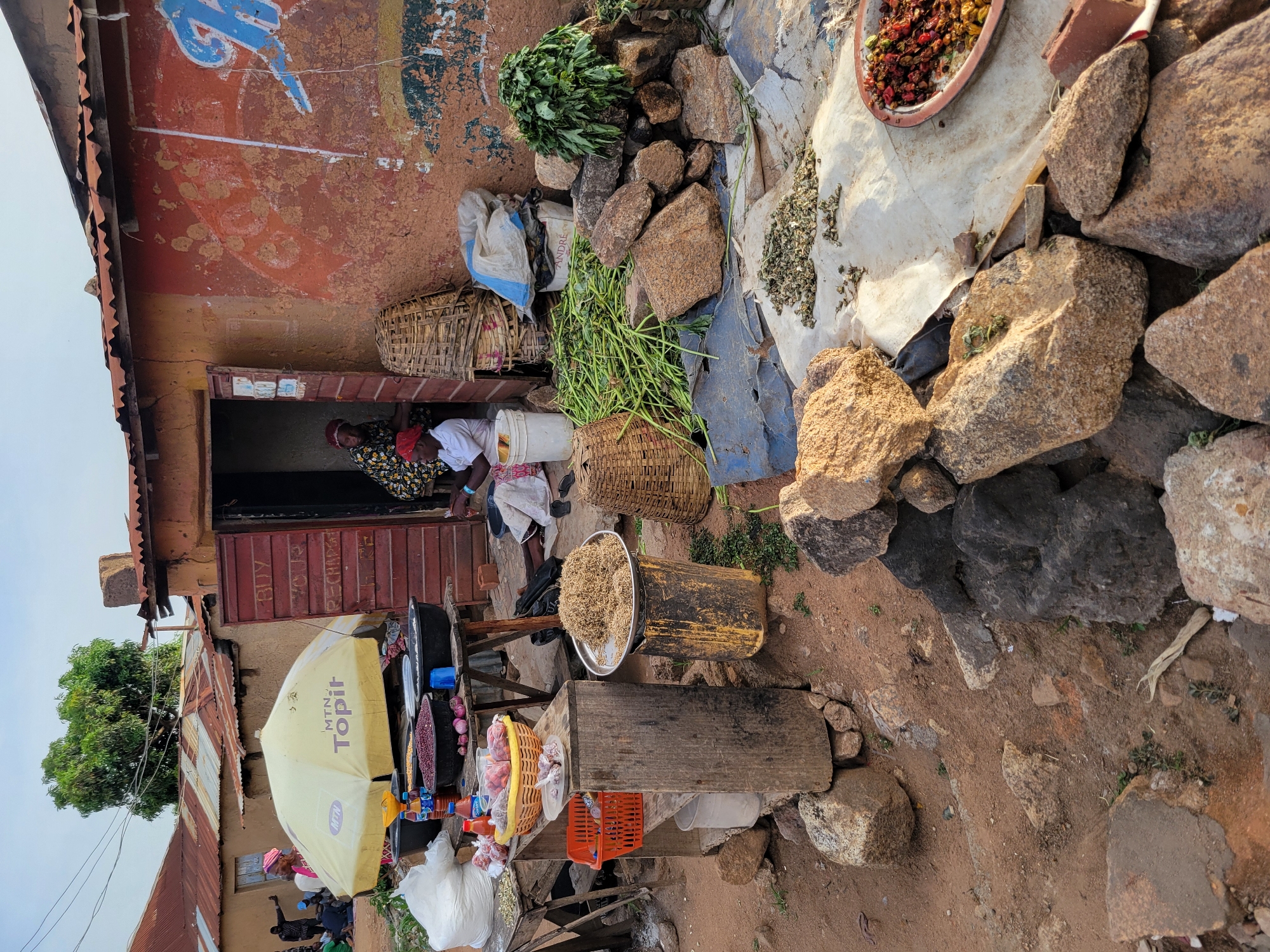 The image depicts a vibrant market scene in Jos, Nigeria. Two women are sitting inside a building. A display of goods is set up outside, including baskets, herbs, and a scale with dried items. A MTN Topil umbrella shades a table laden with assorted items for sale. Rocks and building materials surround the vendor's setup, while various containers and sacks are present. The backdrop features weathered building walls with faded paint.