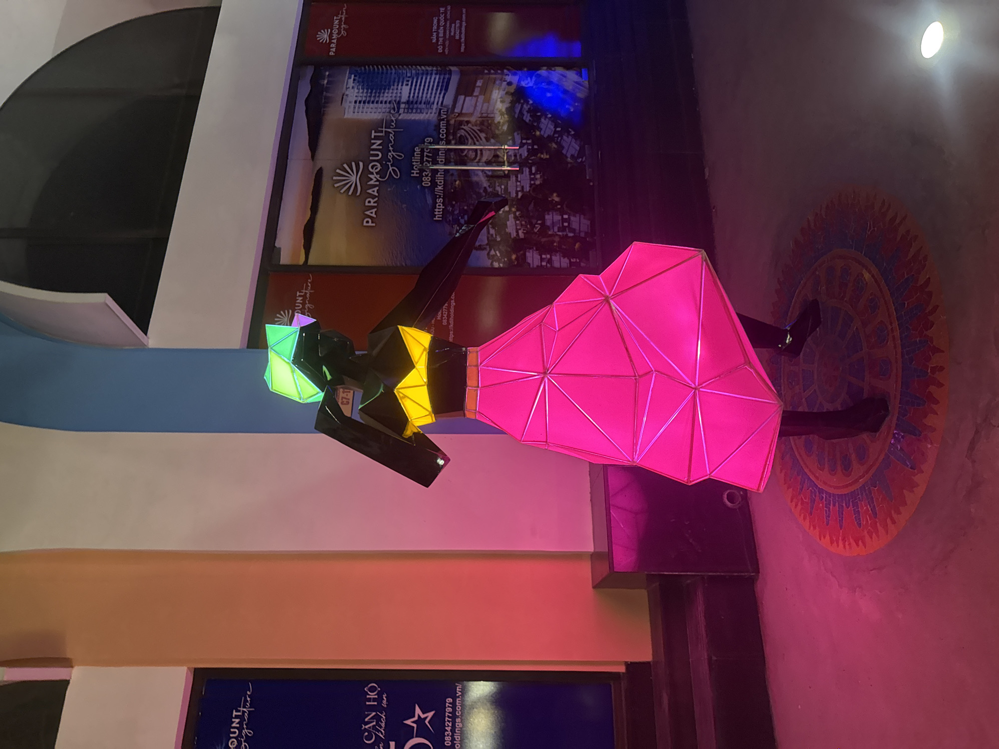 In the city of Nha Trang, specifically in Bac Nha Trang Ward, a vibrant outdoor scene unfolds in front of a building facade. The focal point is a life-sized, illuminated statue of a person dressed in a striking, angular, neon pink skirt and a yellow top. The statue's headwear is a geometric, glowing green structure. It stands on a circular, mosaic-like mat with intricate patterns. The background features posters of what appears to be a resort or residential development, displaying the name "PARAMOUNT Signature" and contact information. The building's architecture includes a light blue curved section above the posters and a textured cream-colored column. The ground is paved and illuminated by the statue's glow and other ambient lighting, casting colorful reflections. The overall atmosphere is festive and modern, suggesting a public space or promotional area.