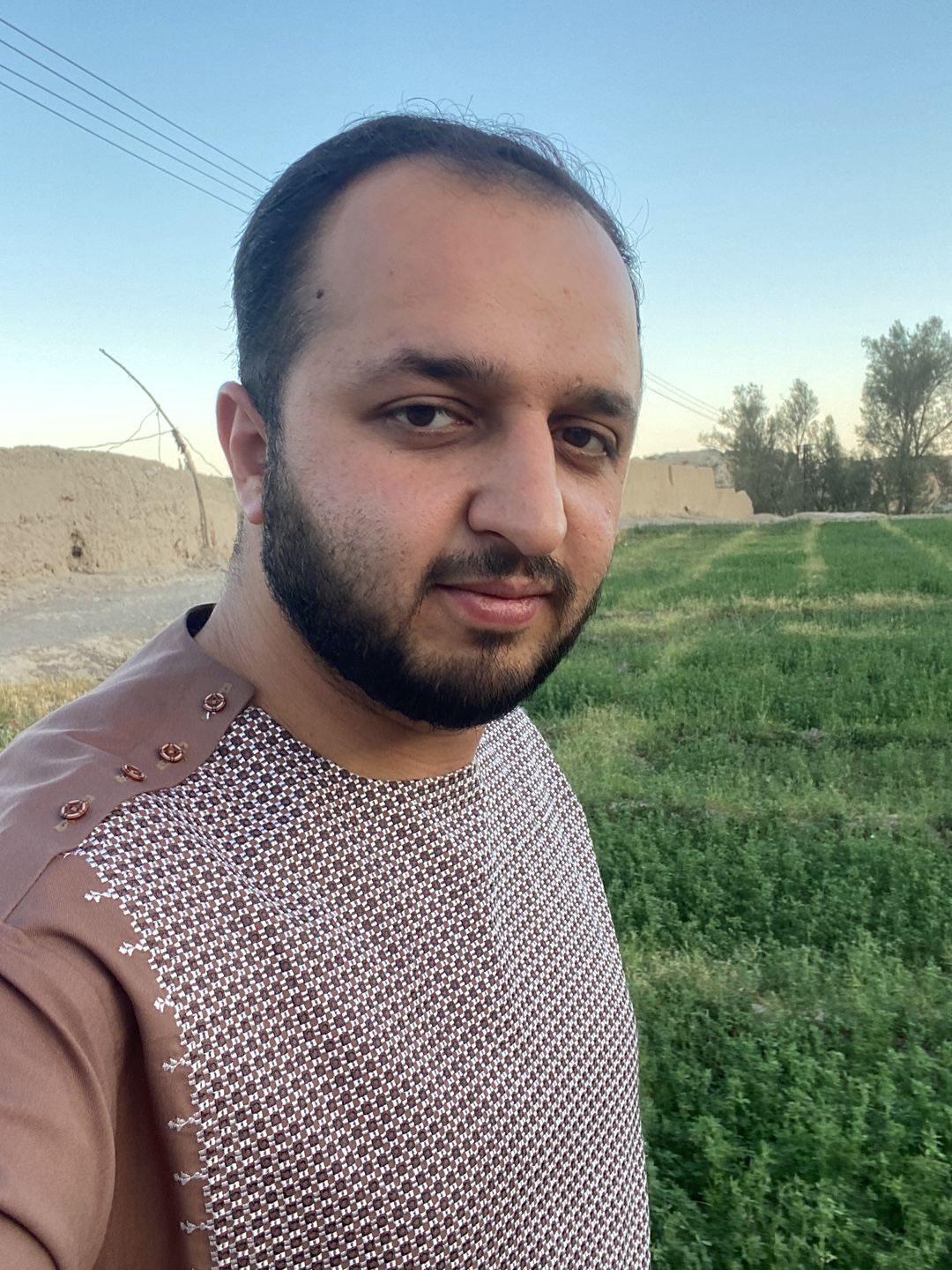A male individual is depicted in a close-up portrait, facing forward and looking directly at the viewer. He has a dark beard and dark hair. He is wearing a long-sleeved brown tunic featuring a repeating white and dark brown checkered pattern across the chest. The right shoulder area of the tunic is solid brown, adorned with three round, reddish-brown decorative buttons and white embroidered details. The scene is an outdoor setting in Kandahar, Afghanistan, under a clear blue sky. To the subject's left, an earthen wall structure is visible, along with multiple overhead power lines and a bare branch. To the subject's right, a field of green vegetation with visible rows extends into the background, where additional earthen structures and a line of trees are present.