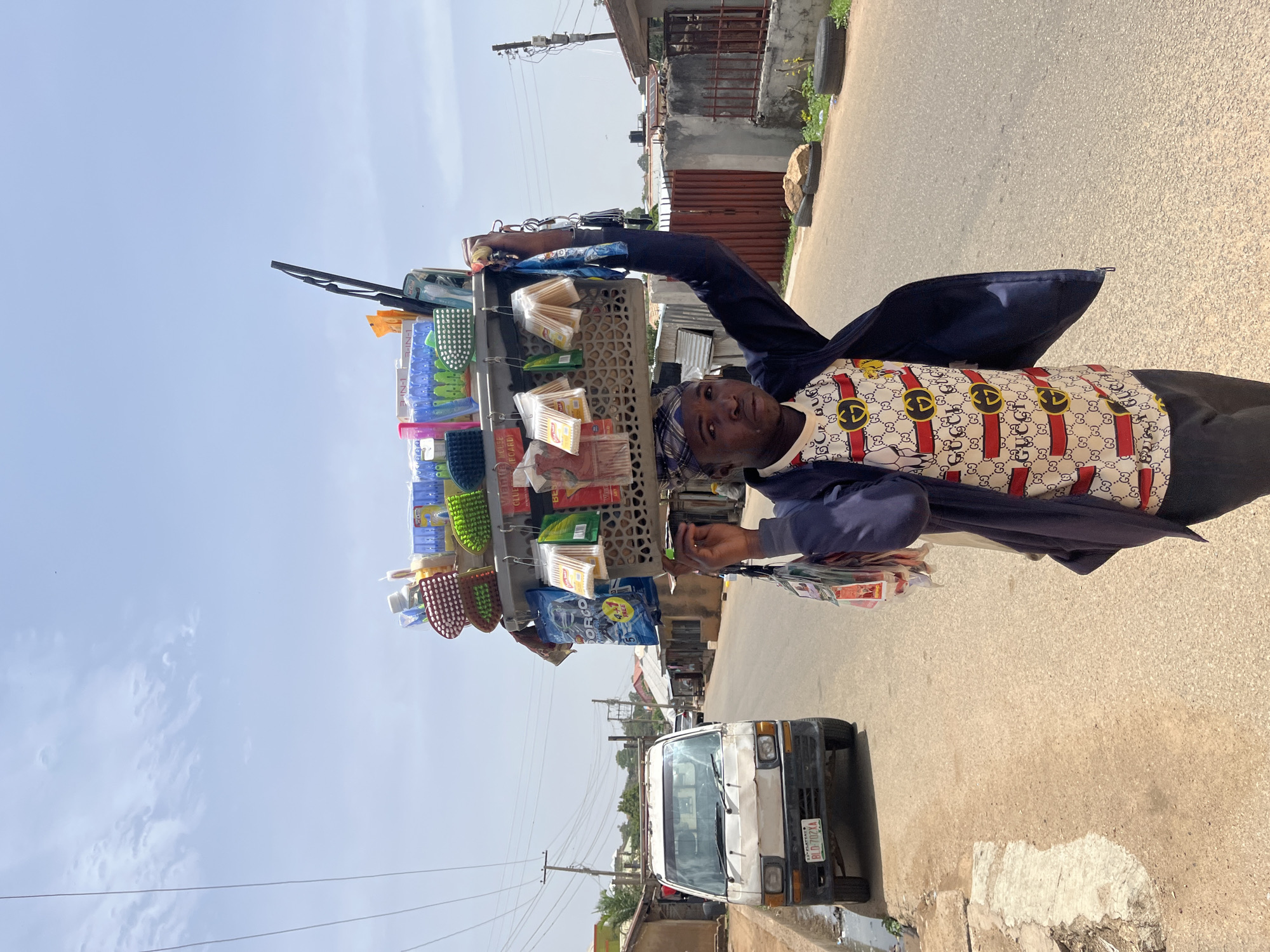 A street vendor stands on a paved road, facing the camera. He has a collection of household items and cleaning supplies displayed on a makeshift rack. He is wearing a patterned shirt under a dark blue jacket, and a bandanna. In the background is a white van. The location is Gura Top, Nigeria.