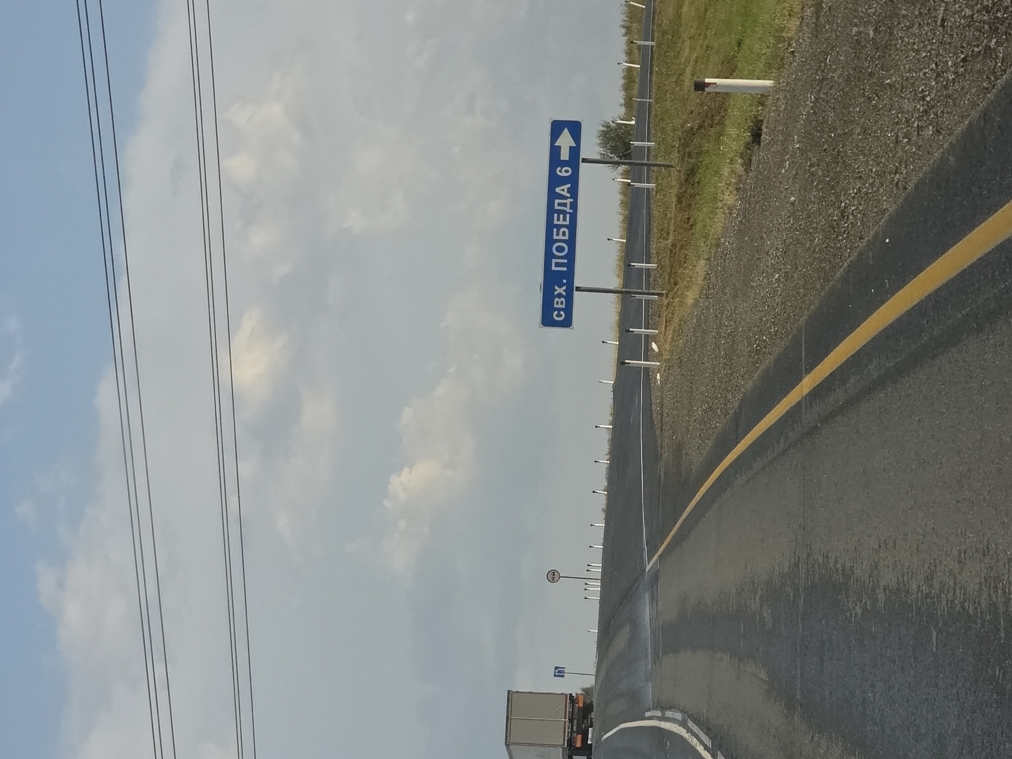 The image shows a road in Troitsk urban district, Russia. A blue sign with white lettering points to the right and reads "свх. ПОБЕДА 6" which translates to "state farm Pobeda 6". There is a truck in the distance to the left of the image. The road is paved and has a yellow line marking the lane. There are roadside markers along the right side of the road. The sky is cloudy with patches of blue sky visible. The environment appears to be rural or semi-rural.