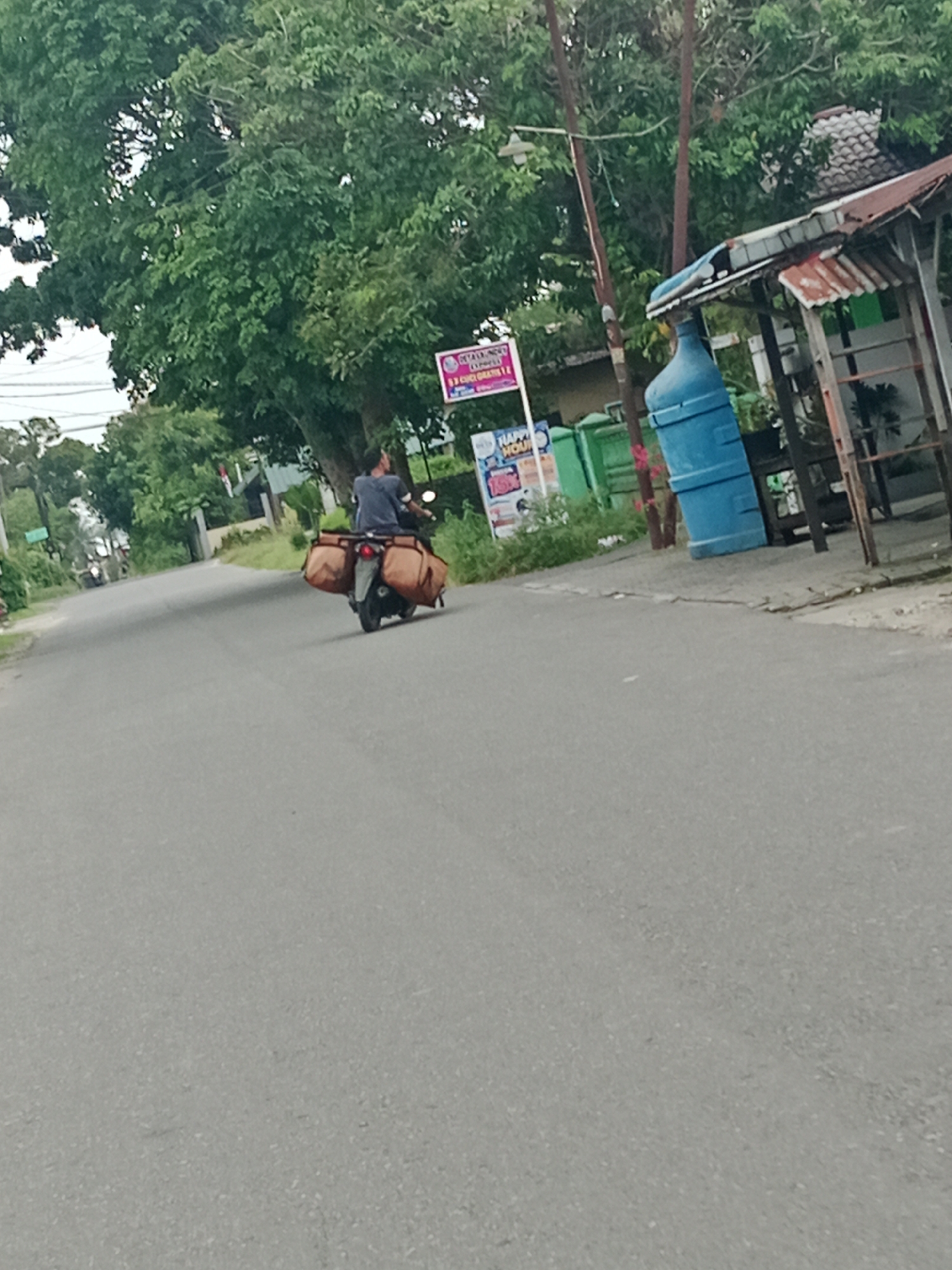The image depicts a road in the City of Binjai, Indonesia, lined with trees and buildings. A man is riding a motorcycle, carrying two large brown saddlebags on the rear. The motorcycle is moving away from the camera on a paved road. To the right of the road, there is a small stall with a corrugated iron roof and a large blue water dispenser. A sign with Indonesian text is visible in the background. The sky is overcast.