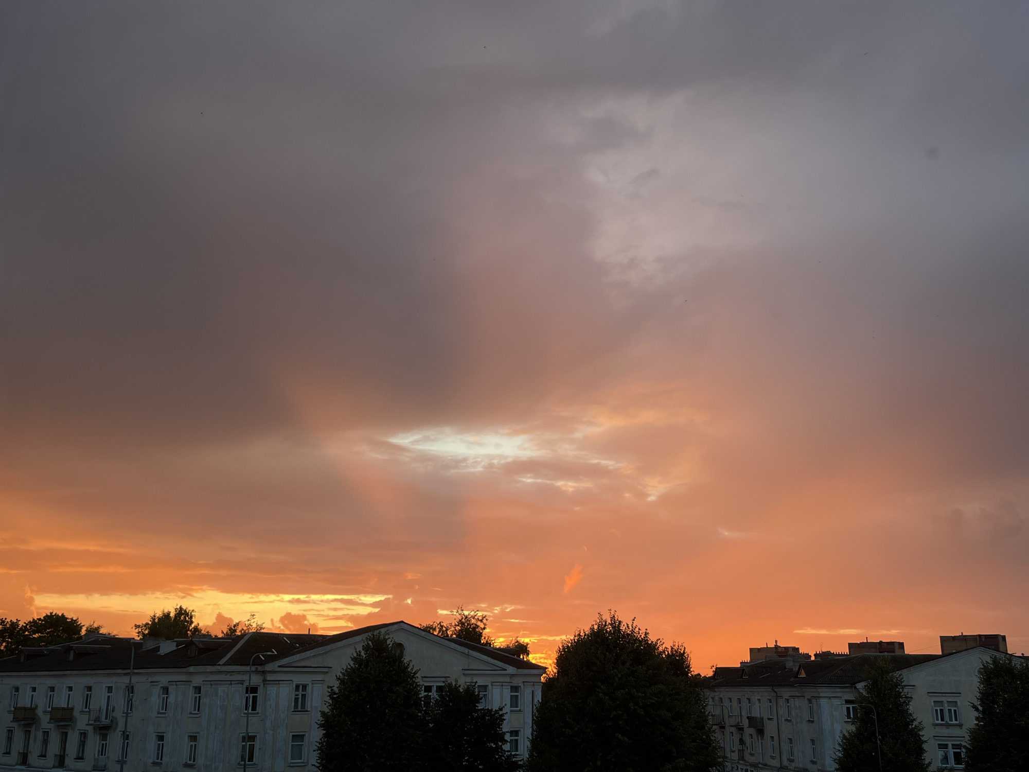 The image shows a dramatic sunset over the city of Kohtla-Järve linn, Estonia. The sky is a gradient of colors, from deep purple and gray at the top to fiery orange and yellow near the horizon. Rays of sunlight pierce through the clouds, creating a spotlight effect in the center of the sky. In the foreground, the rooftops of several apartment buildings are visible, along with silhouetted trees. The buildings appear to be Soviet-era architecture, characterized by their long, rectangular shapes and numerous windows. The overall impression is one of a beautiful and somewhat melancholic end to the day. There are no people or visible actions depicted in the image.