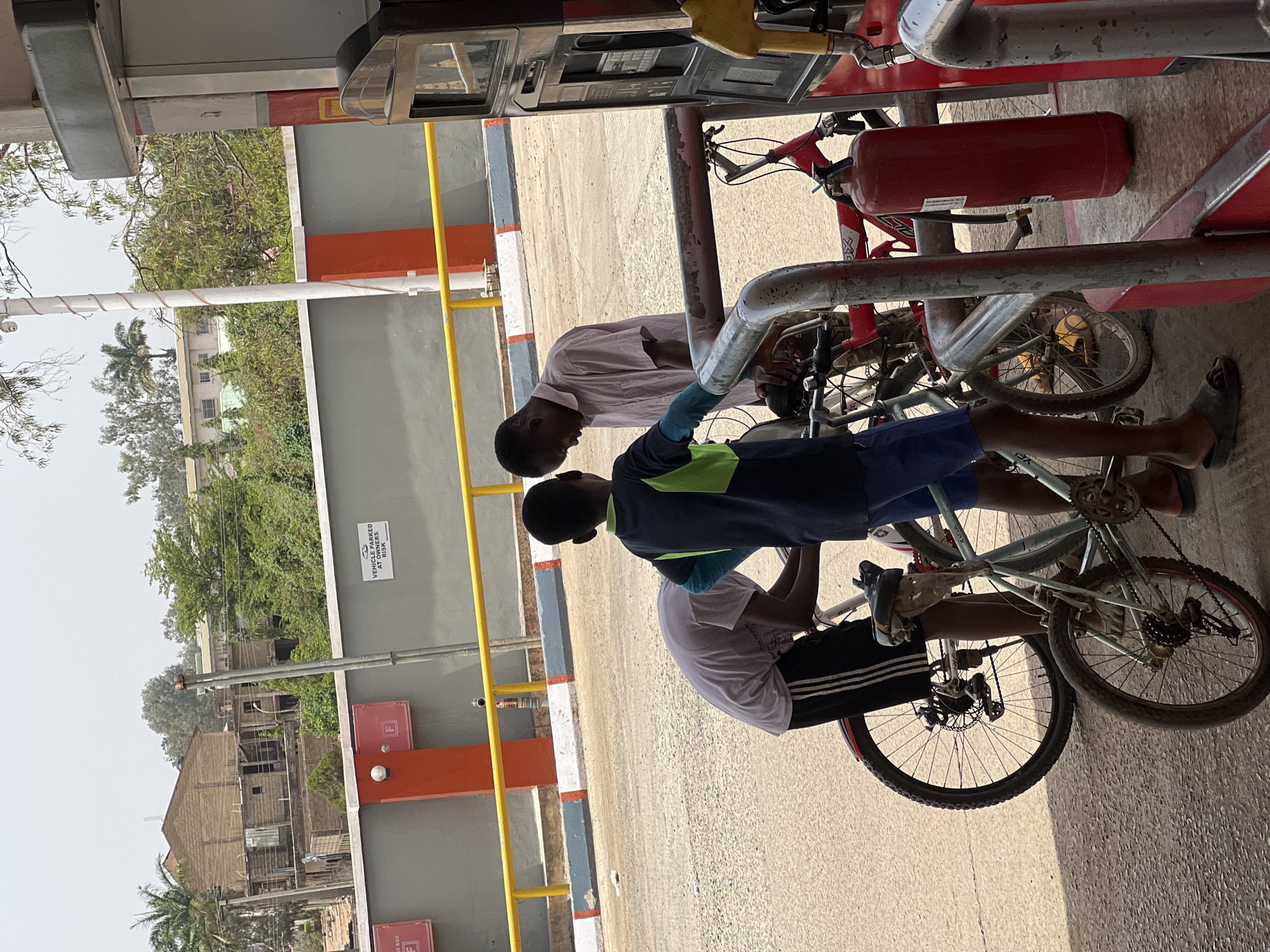 Two individuals, one in a white shirt and the other in a dark shirt with blue and green accents, are interacting near bicycles. The location appears to be a roadside area with a building and trees in the background. Based on the presence of a fuel pump, it is likely a gas station in Jos, Nigeria.