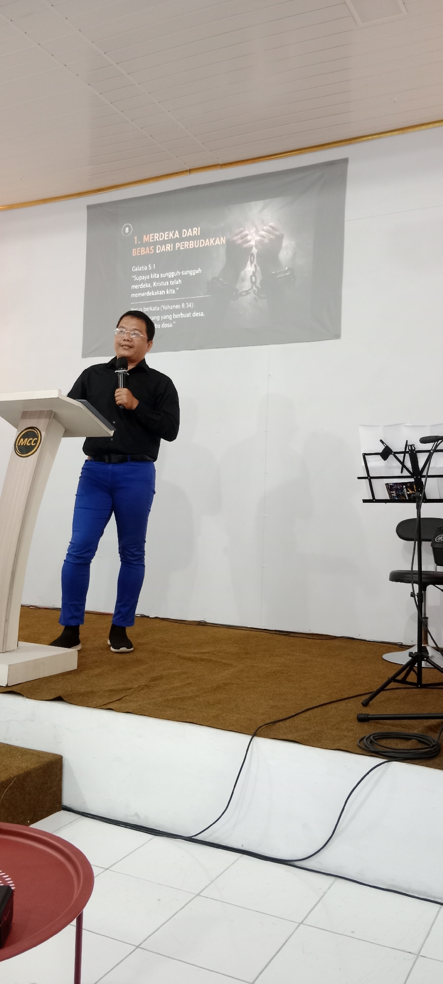 A man wearing glasses, a black long-sleeved shirt, and bright blue pants stands behind a wooden podium labeled "MCC". He holds a microphone in his right hand and appears to be speaking. Behind him, a projection screen displays text in Indonesian, with the title "1. MERDEKA DARI BEBAS DARI PERBUDAKAN" (1. FREEDOM FROM SLAVERY) prominently featured, along with biblical verses. To the right of the podium, a keyboard stand with a stool is visible. The room has white walls and a white ceiling with visible paneling. The floor is covered with a brown carpet in the foreground, transitioning to white tiles in the background. The overall scene suggests an indoor event, possibly a church service or conference, in a location like Magelang, Indonesia.